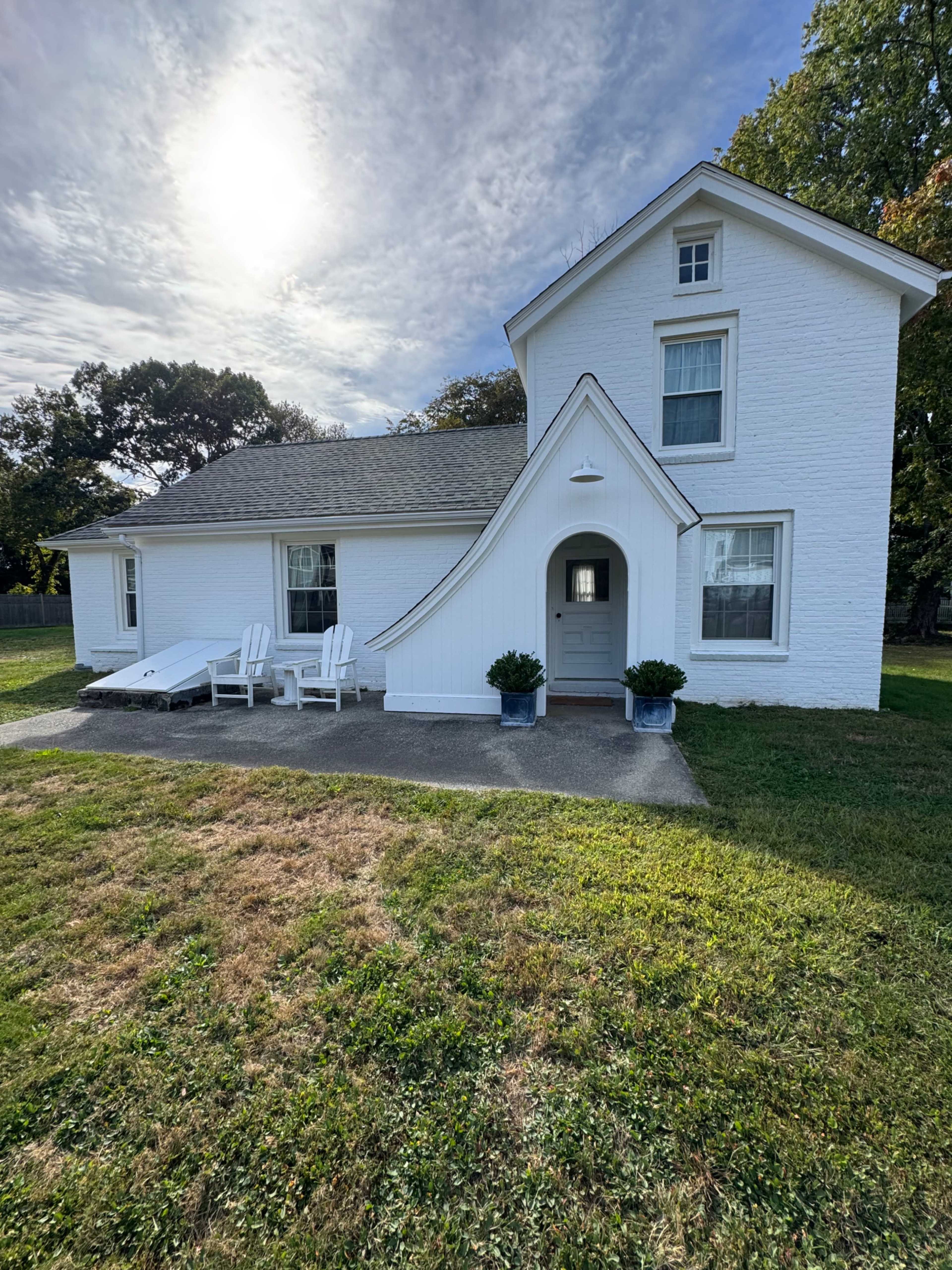 The image shows a white, two-story house with unique architectural features, including a curved entrance, a front yard, and two white adirondack chairs on a concrete patio.