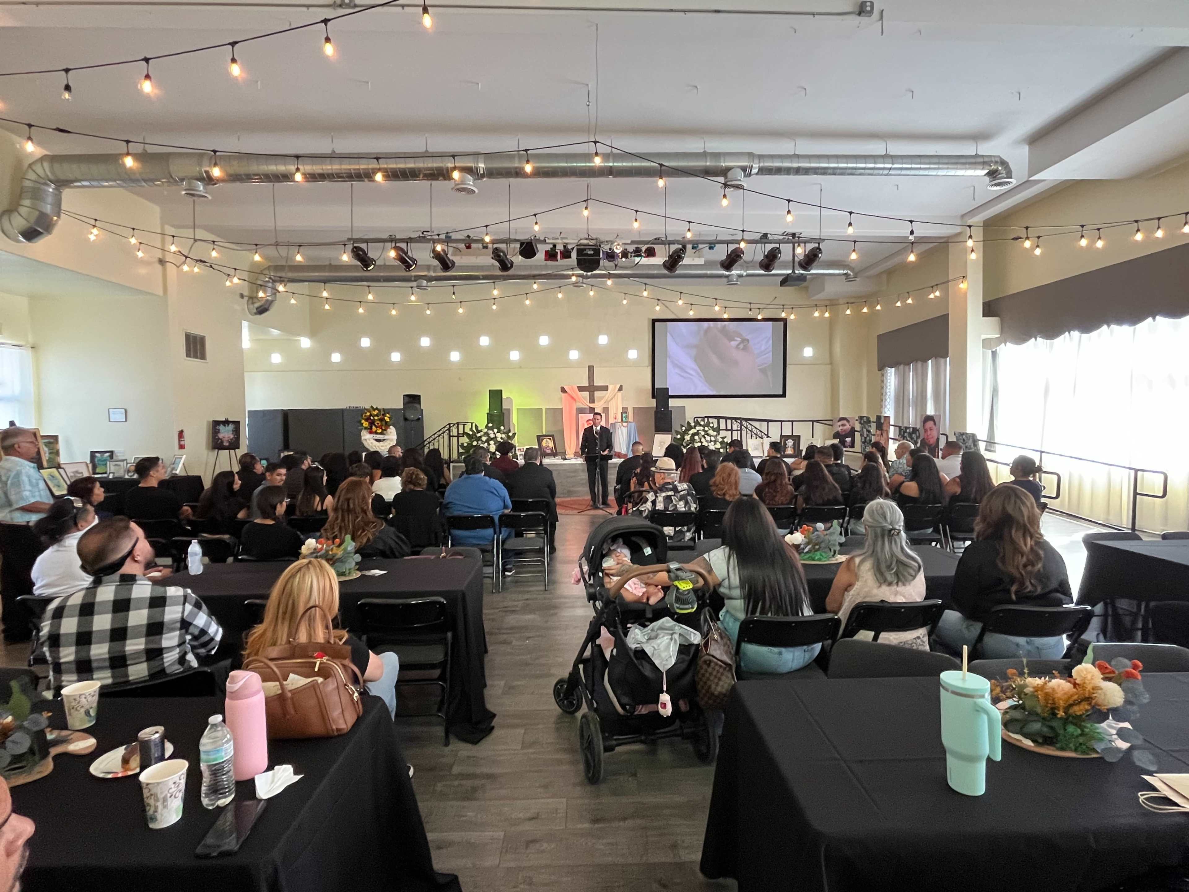 A gathering of people sits at tables in a room decorated with string lights, while a speaker presents on a stage in front of a screen.