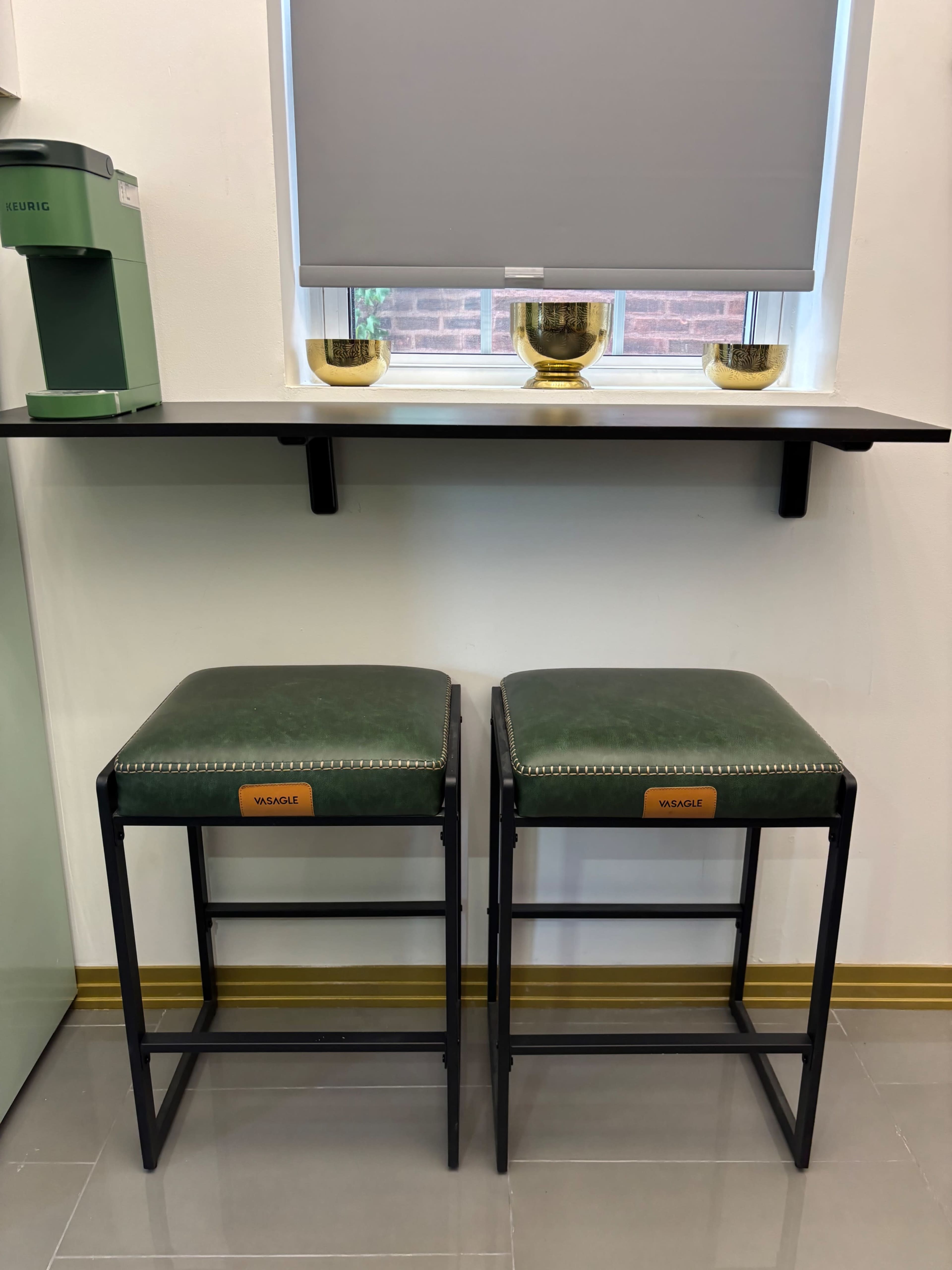 The image shows two green cushioned bar stools with black metal frames positioned under a shelf near a window with a closed blind.