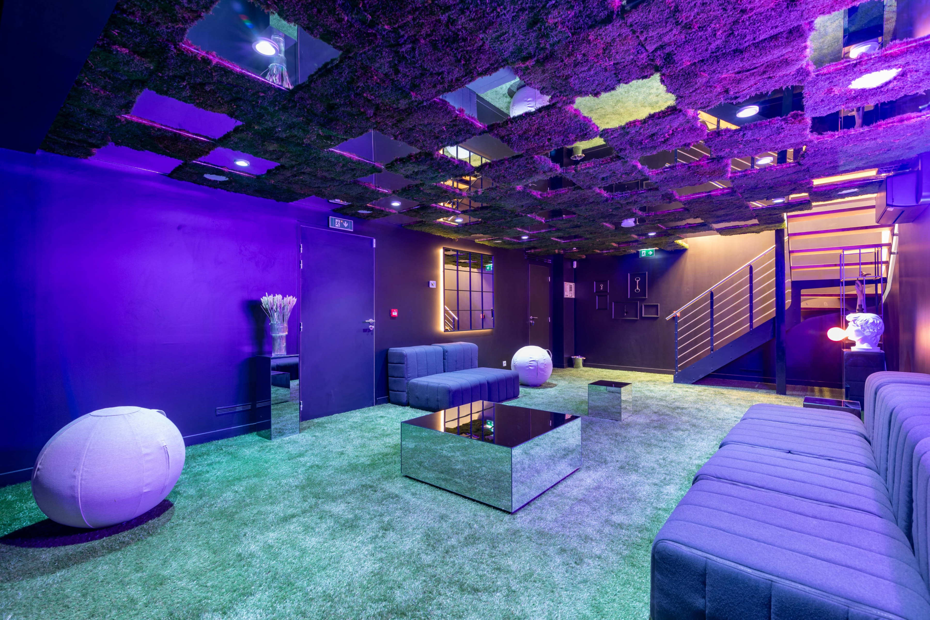 A modern lounge area features green carpeting, mirrored furniture, and a ceiling with reflective panels, illuminated by purple lighting.