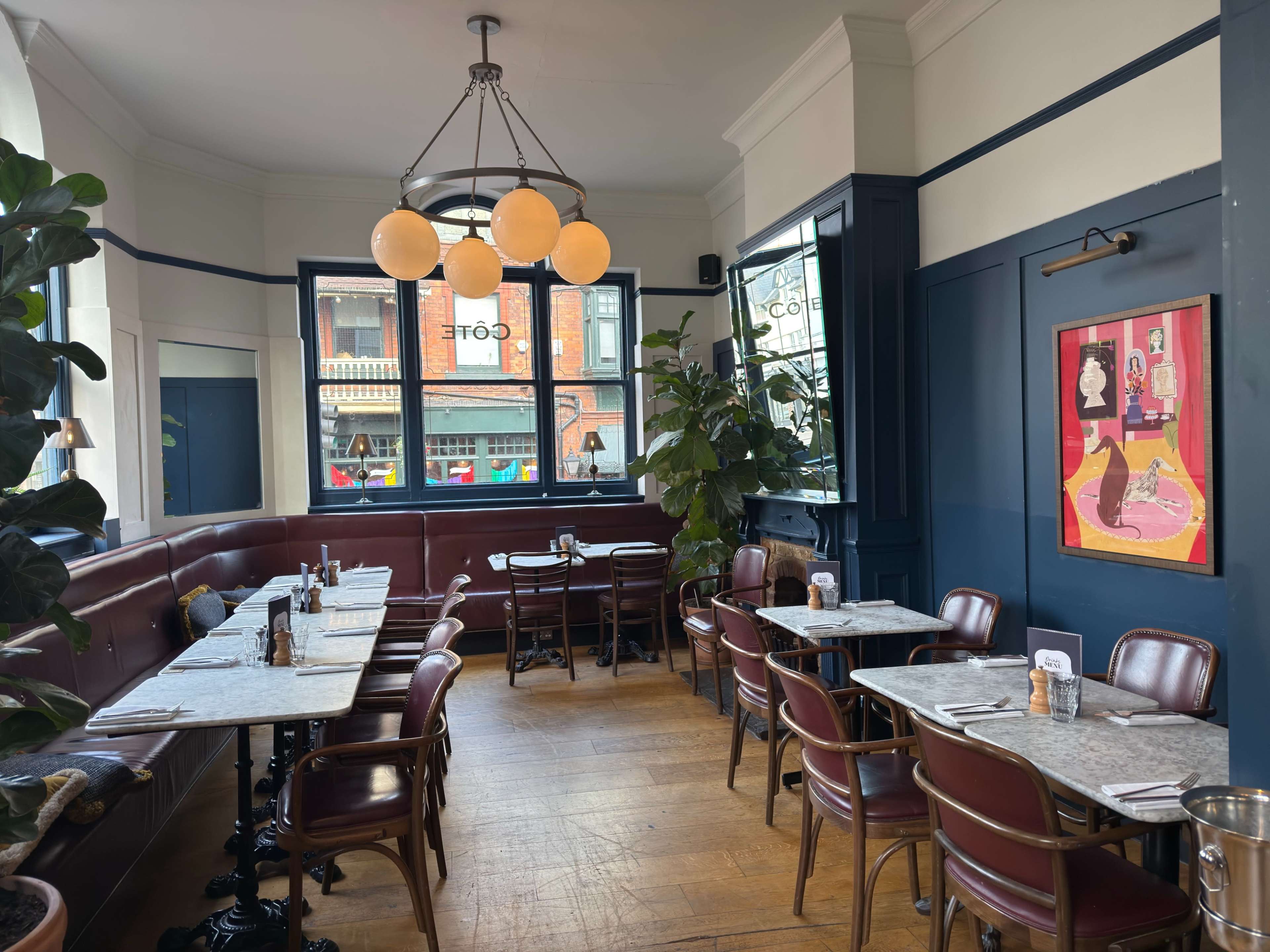 The image shows a cozy restaurant interior with dark blue walls, wooden flooring, and marble-topped tables arranged in a well-lit space, featuring a large window and decorative plants.