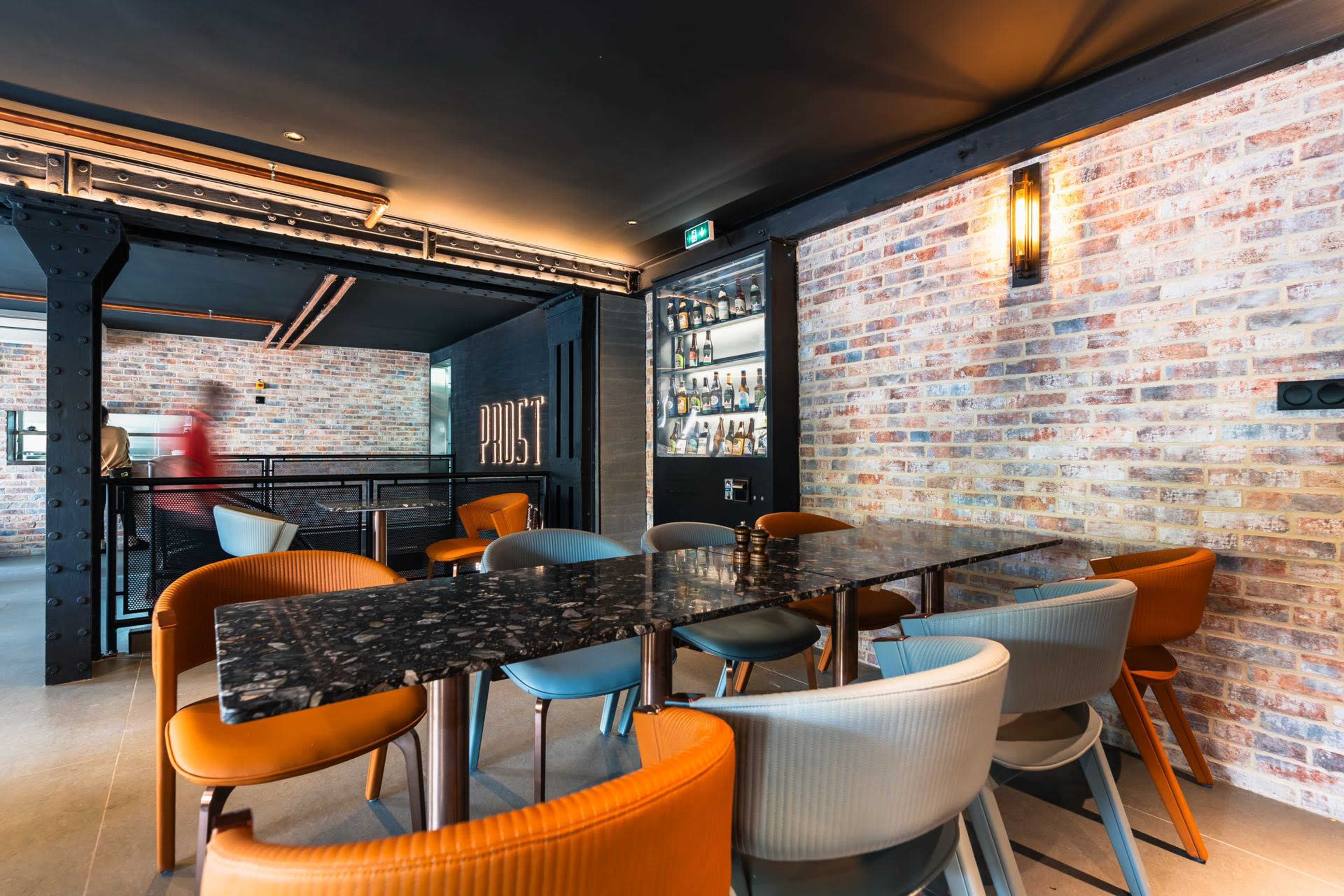 The image shows a modern dining area with a long black table surrounded by colorful chairs, set against a brick wall and industrial-style decor.