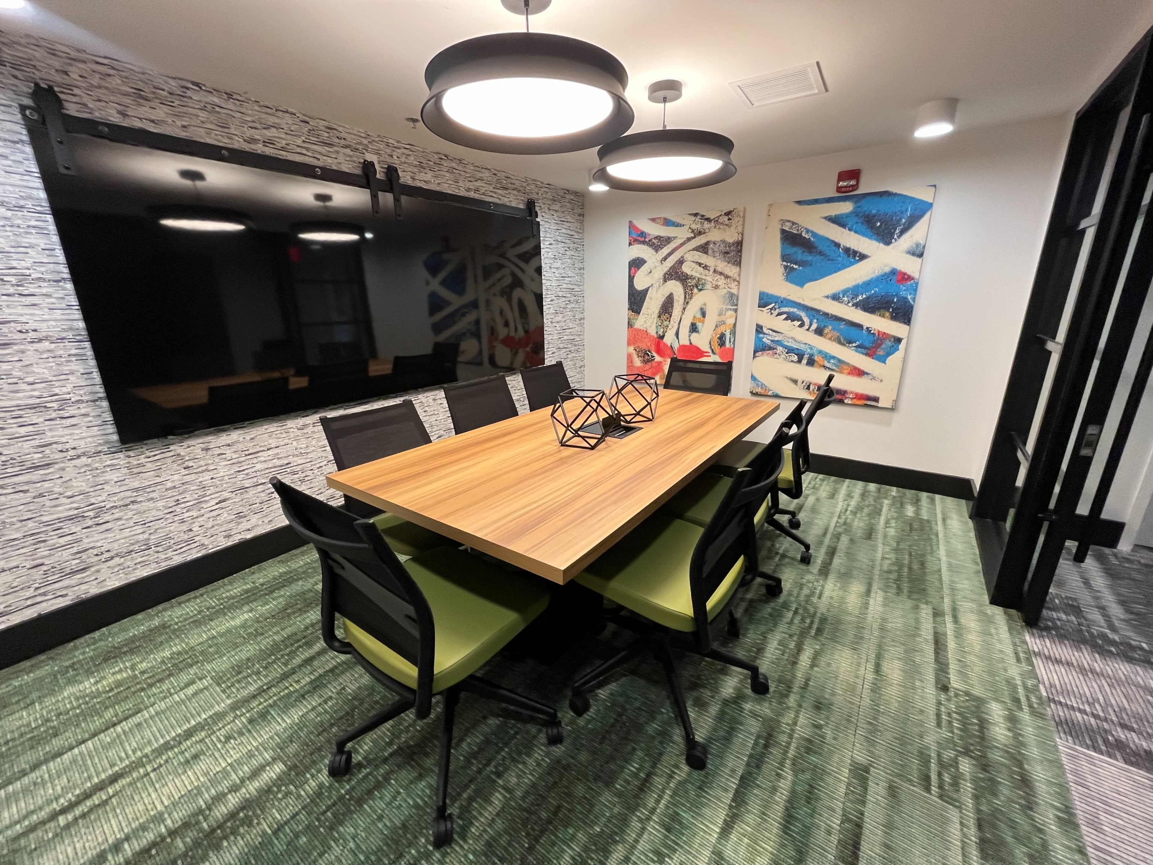 A modern conference room features a long wooden table surrounded by black chairs with green accents, illuminated by pendant lights, and includes a large mirror and abstract artworks on the walls.