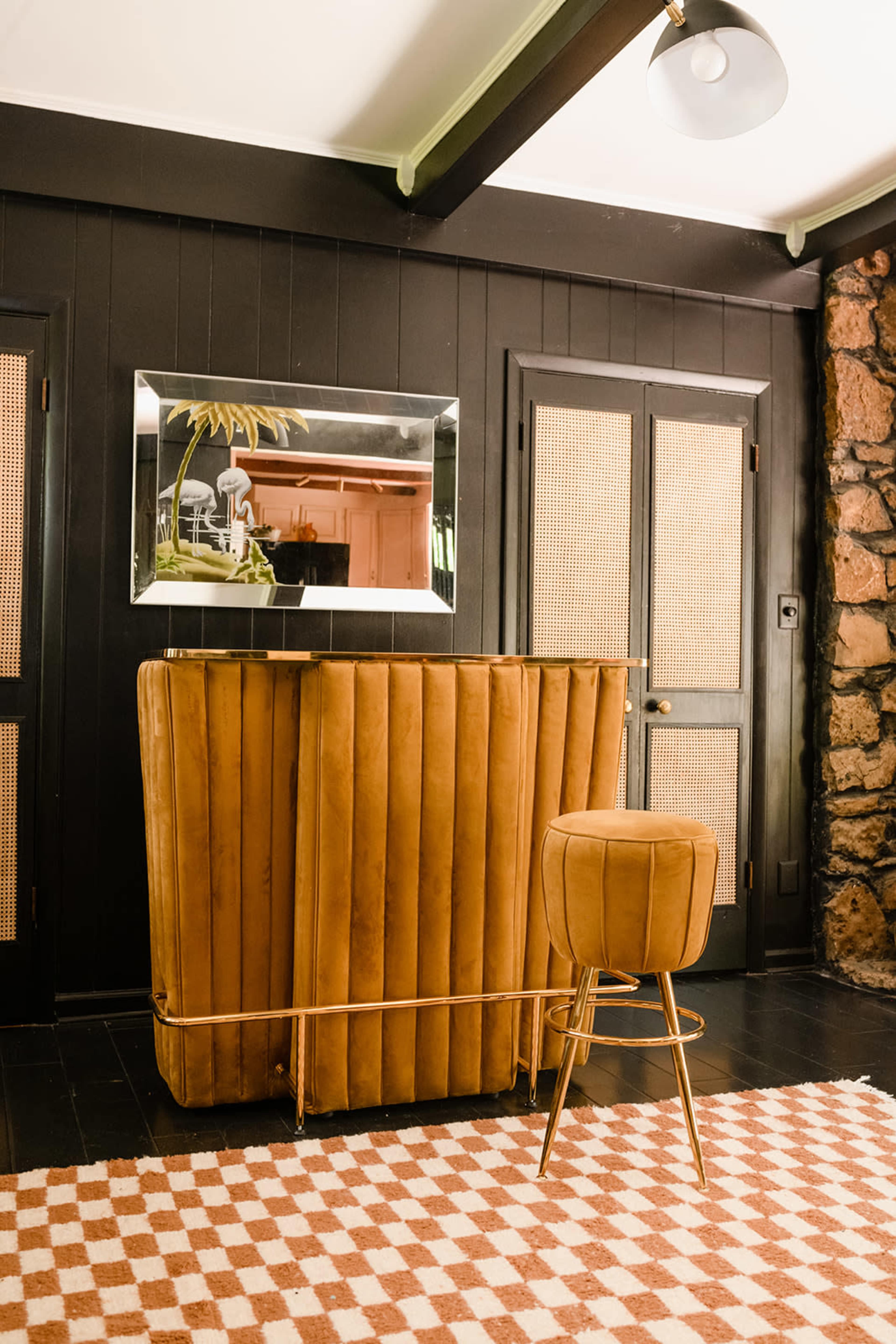 A stylish bar area featuring a brown ribbed leather bar with a matching stool and a mirror above it, set against dark walls and a stone accent wall.
