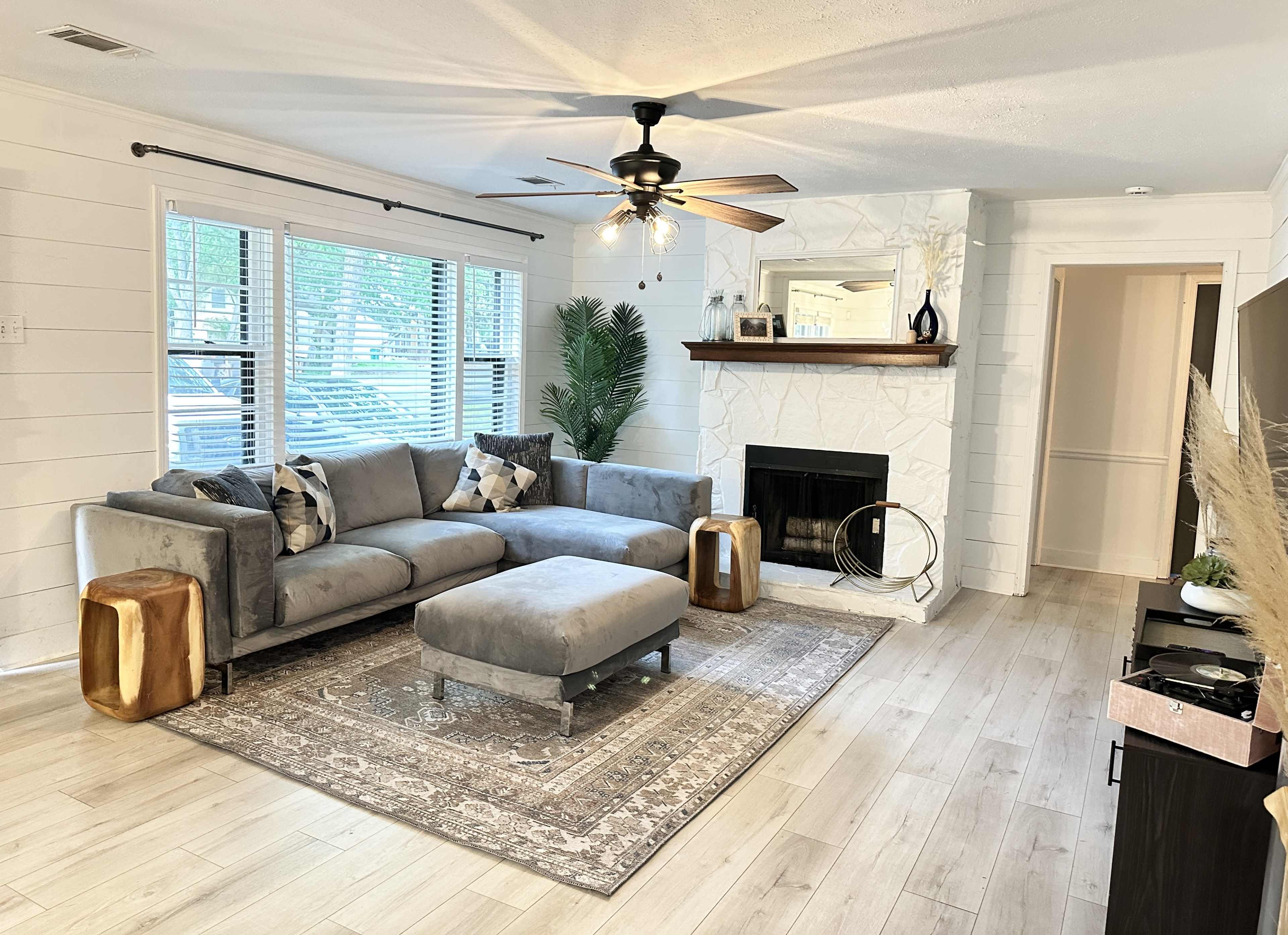 The living room features a gray sectional sofa with decorative pillows, a marble fireplace, and large windows allowing natural light, complemented by a patterned area rug and wooden side tables.