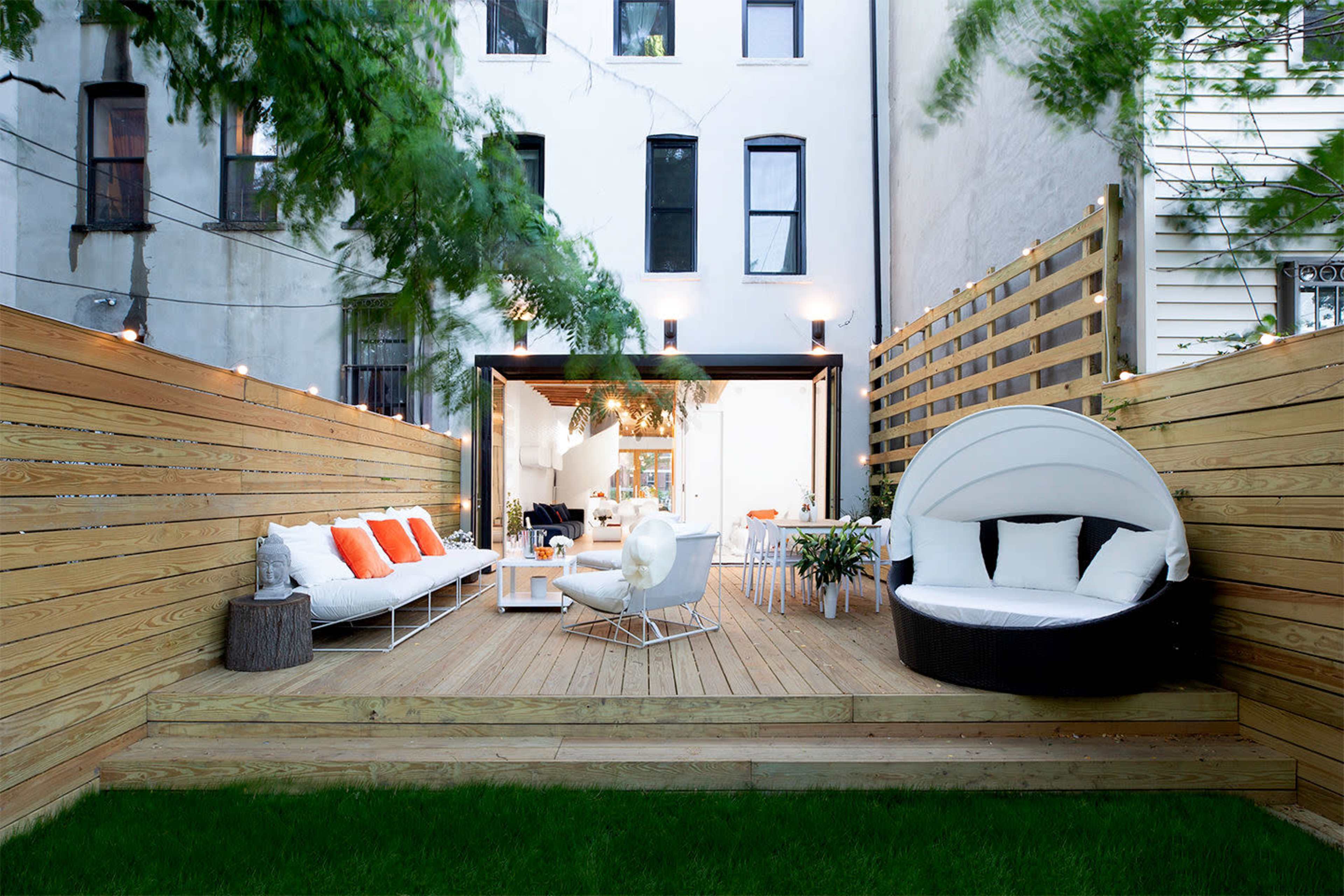 A modern outdoor patio area with wooden decking, featuring a white seating arrangement with orange cushions, a round sunbed, and a dining space adjacent to a spacious house.
