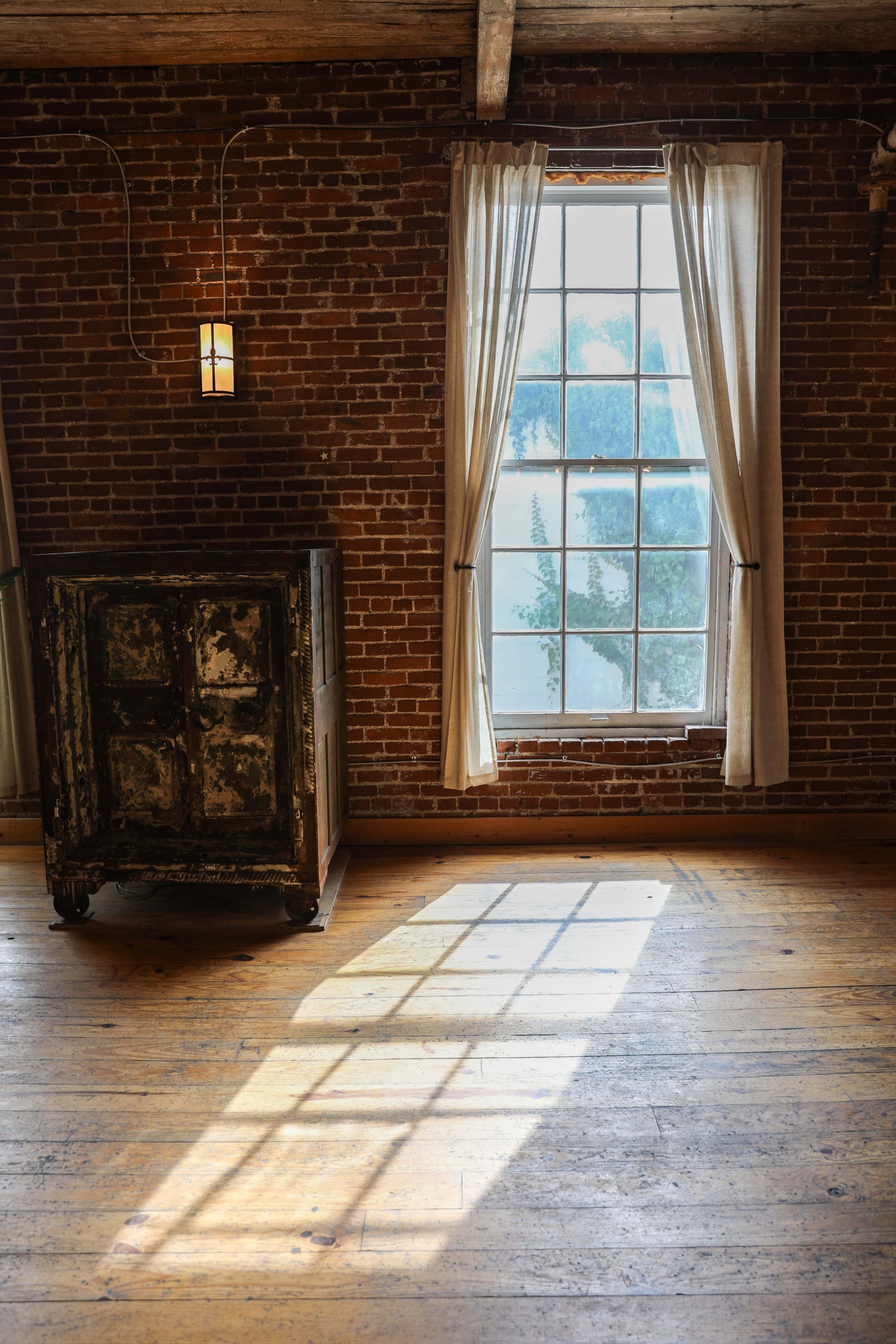 A dimly lit room features a large window with sheer curtains, illuminating the wooden floor and casting a shadow from a vintage cabinet nearby.