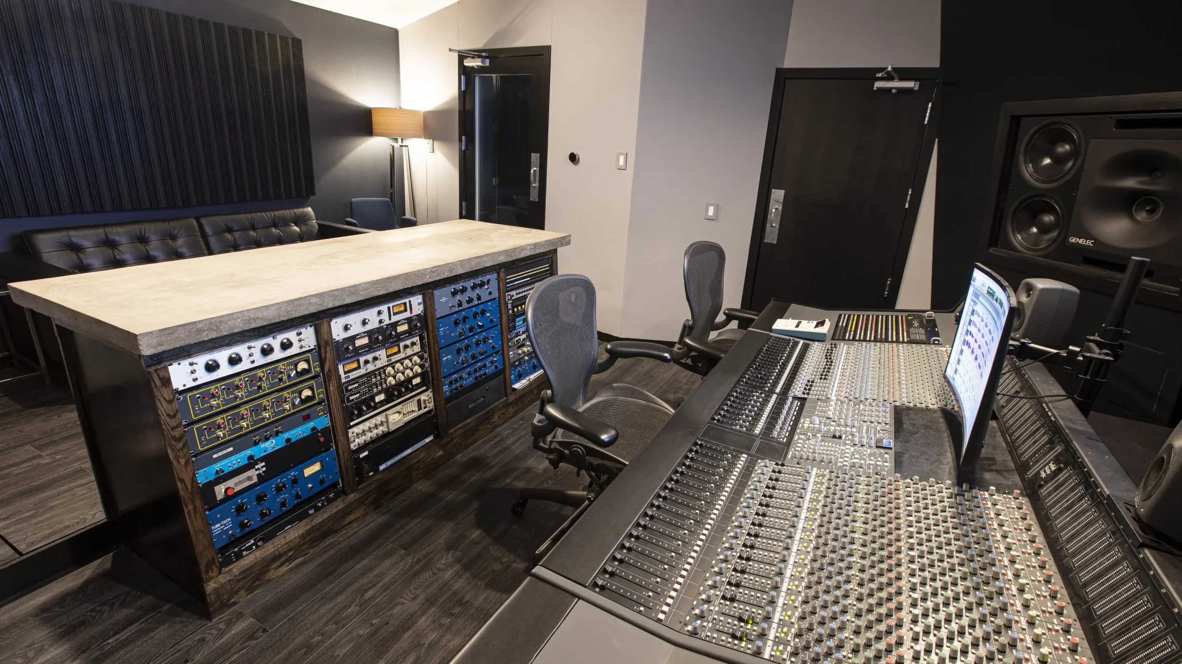 The image shows a modern music studio with a control desk equipped with audio mixing equipment and various rack-mounted gear, as well as a seating area in the background.