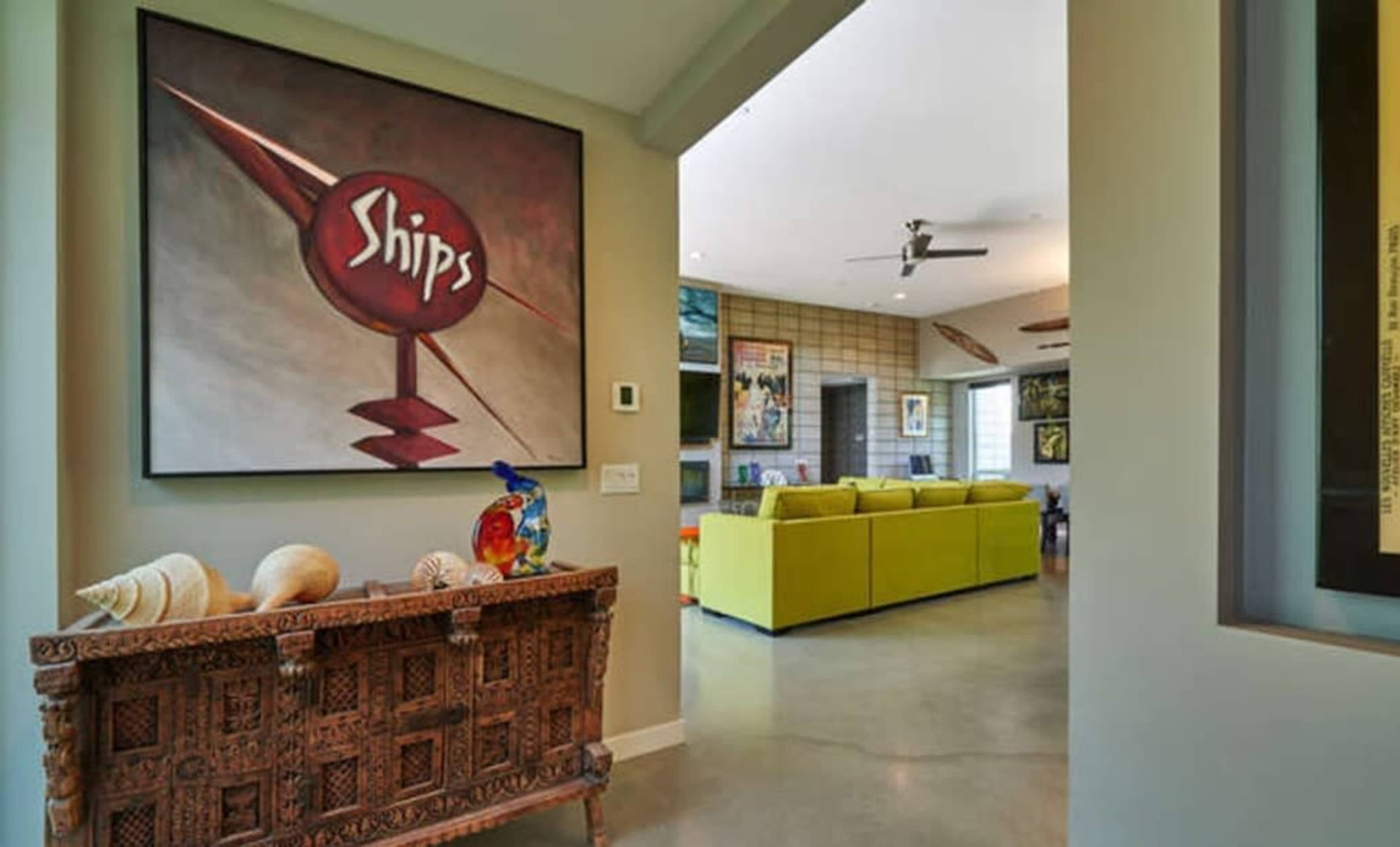 A foyer with a large painting of a red circle that says "Ships" above a decorative wooden console table and a view into a modern living room with green furniture.