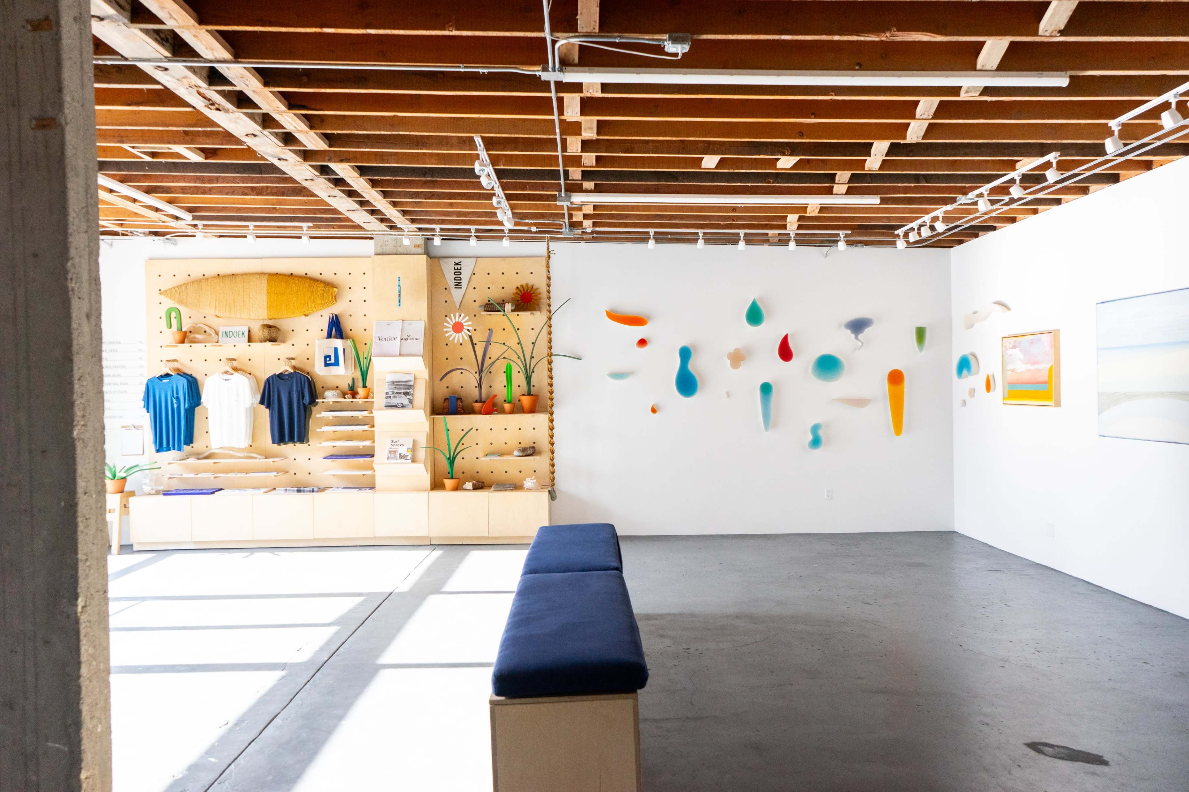 The image shows a contemporary art gallery with a wooden ceiling, featuring a display wall of colorful abstract shapes and a seating area beside a merchandise wall.