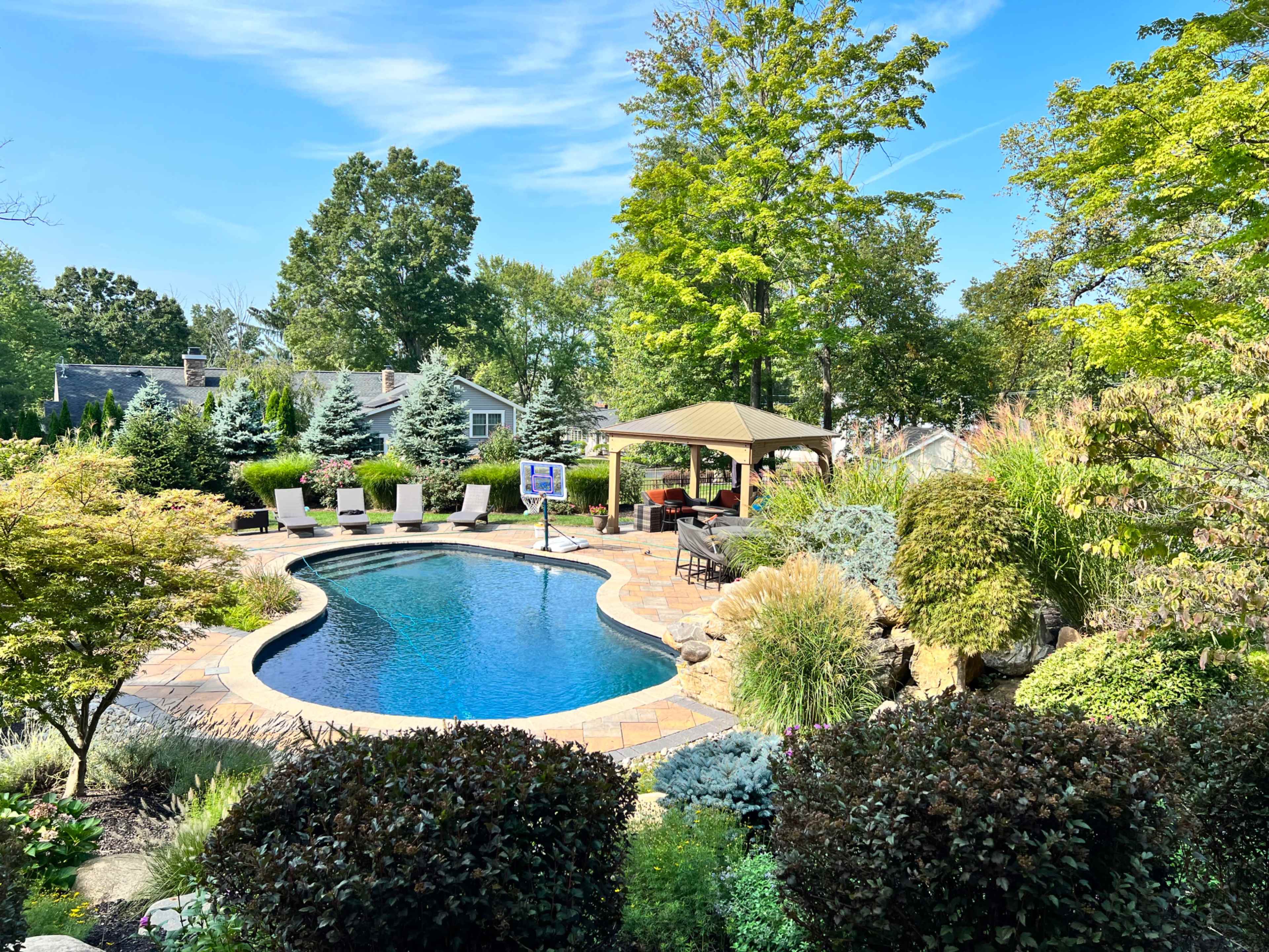 The image shows a landscaped backyard with a swimming pool, surrounded by lush greenery and outdoor seating under a gazebo.