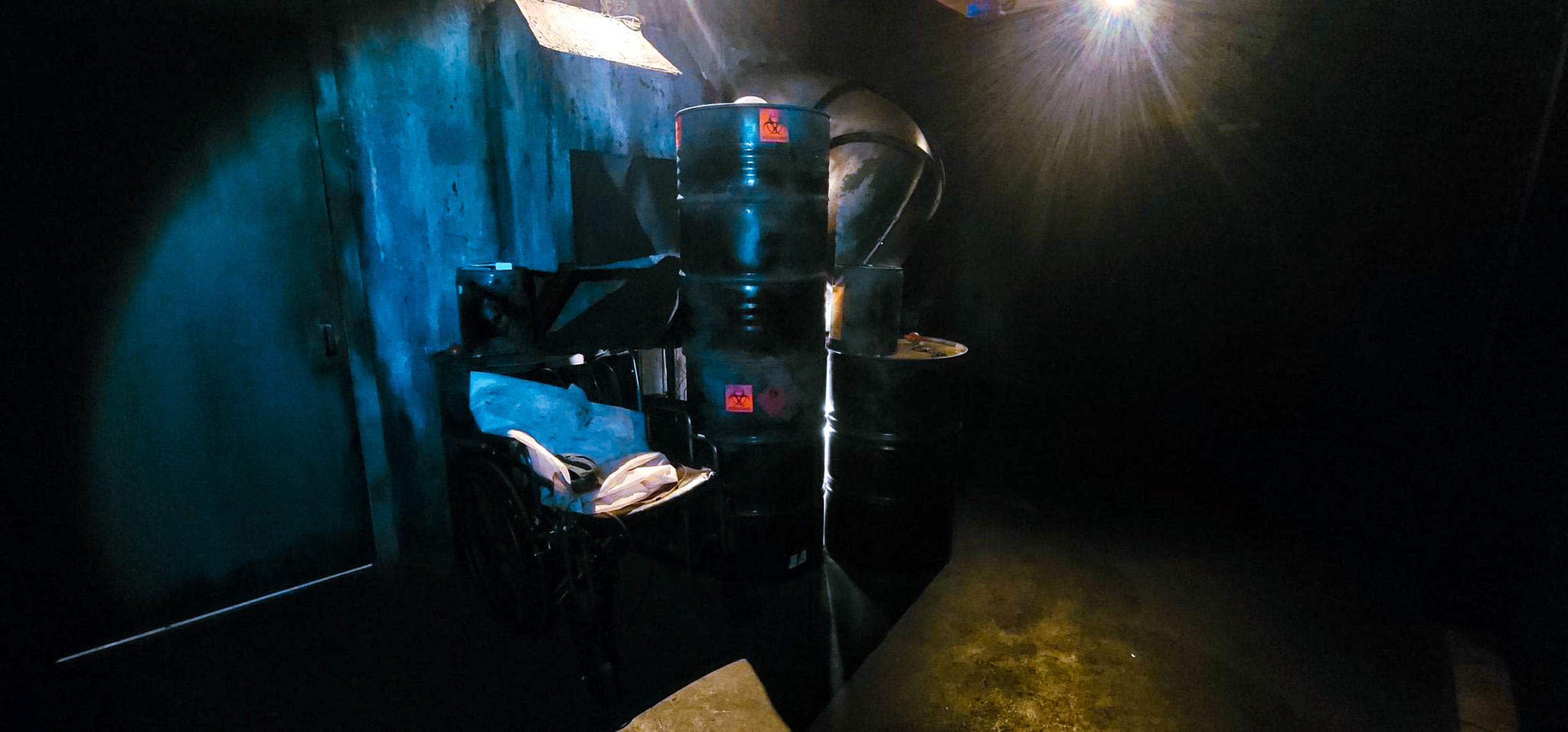 The image shows a dimly lit industrial space featuring several black barrels and a metal workbench against a dark wall.