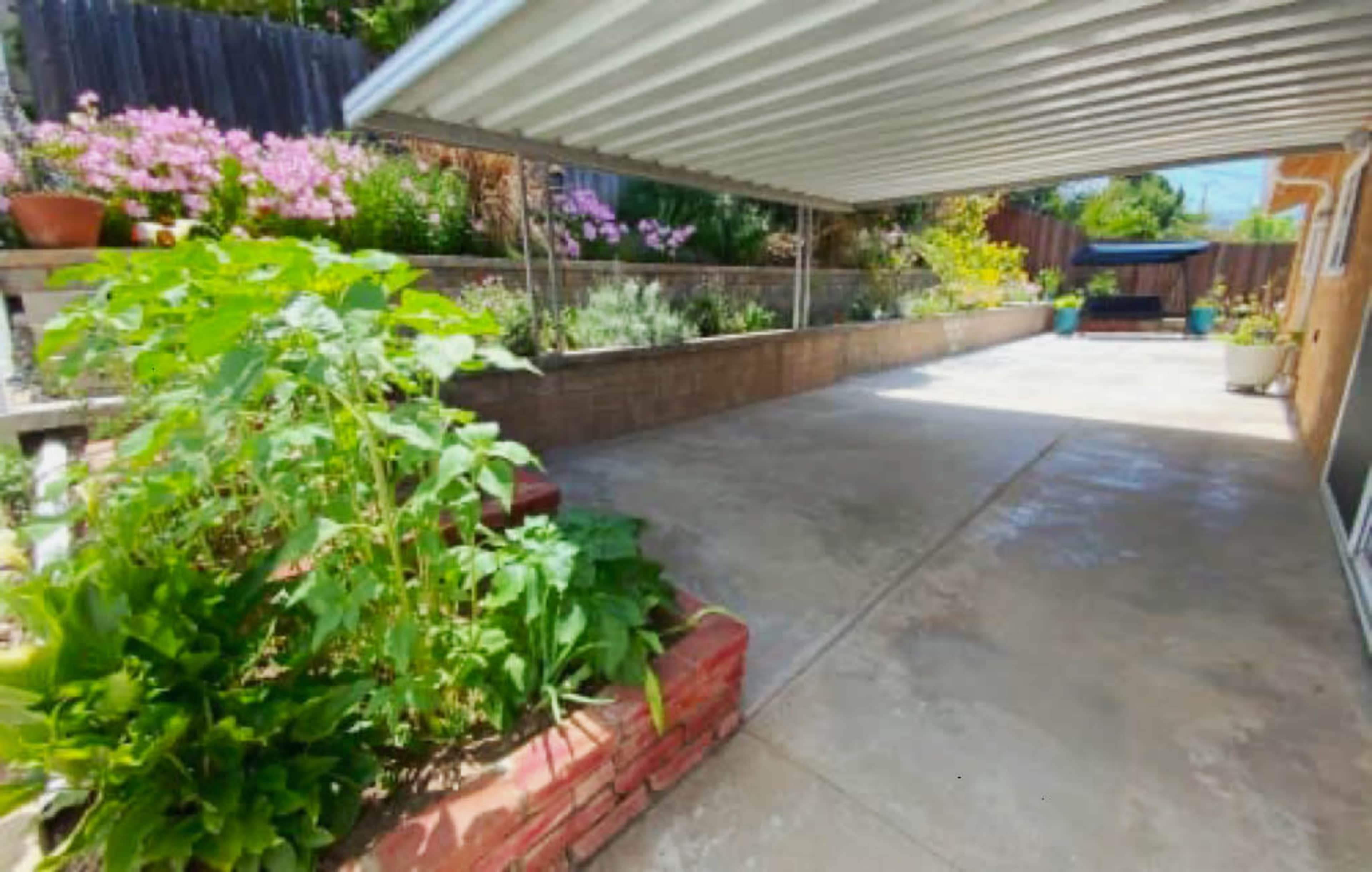 The image shows a concrete patio area under a metal awning, with a garden featuring flowers and plants along one side.
