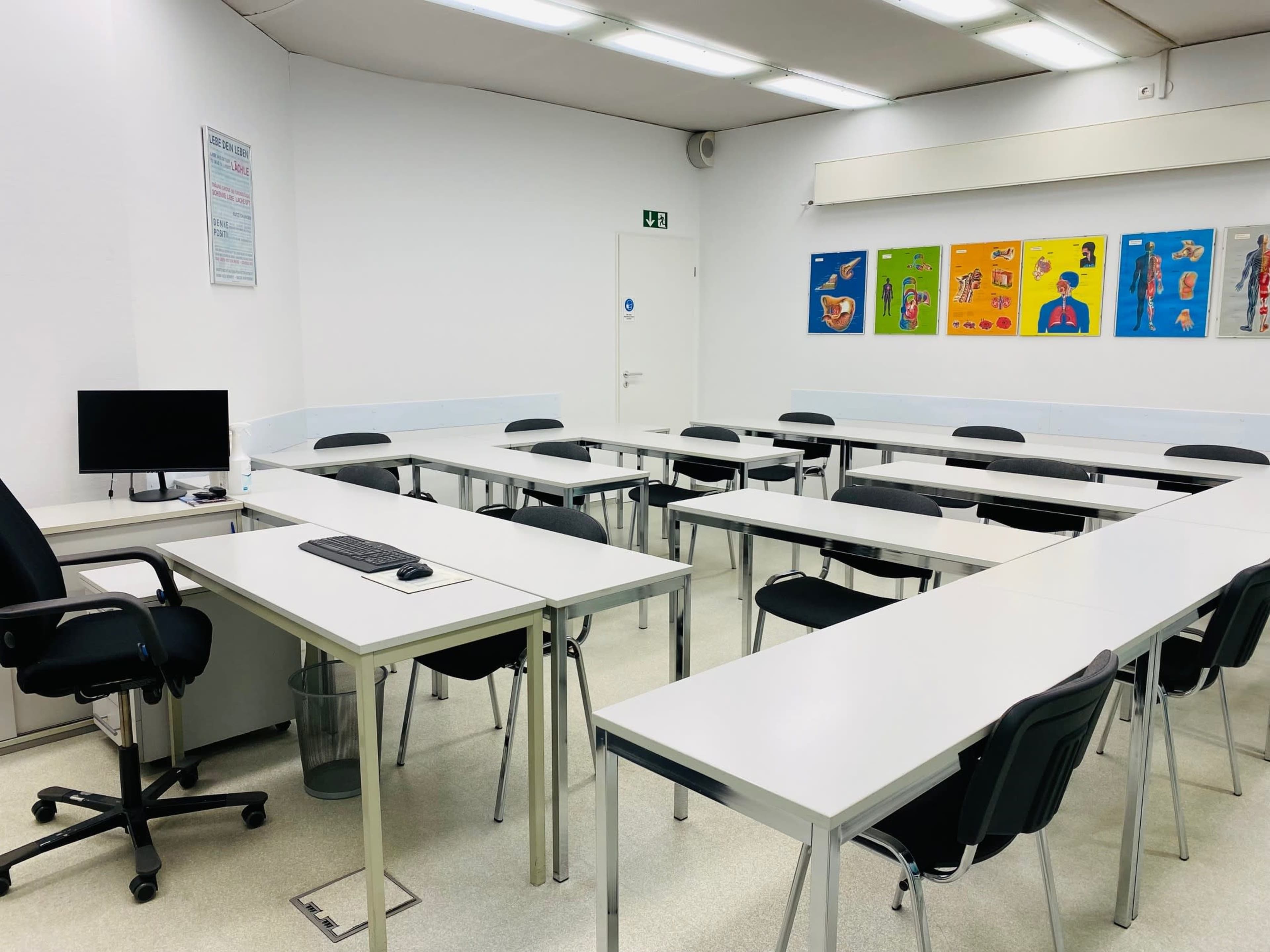 The image shows a classroom with several white desks arranged in a U-shape, a computer on one desk, and colorful educational posters on the wall.