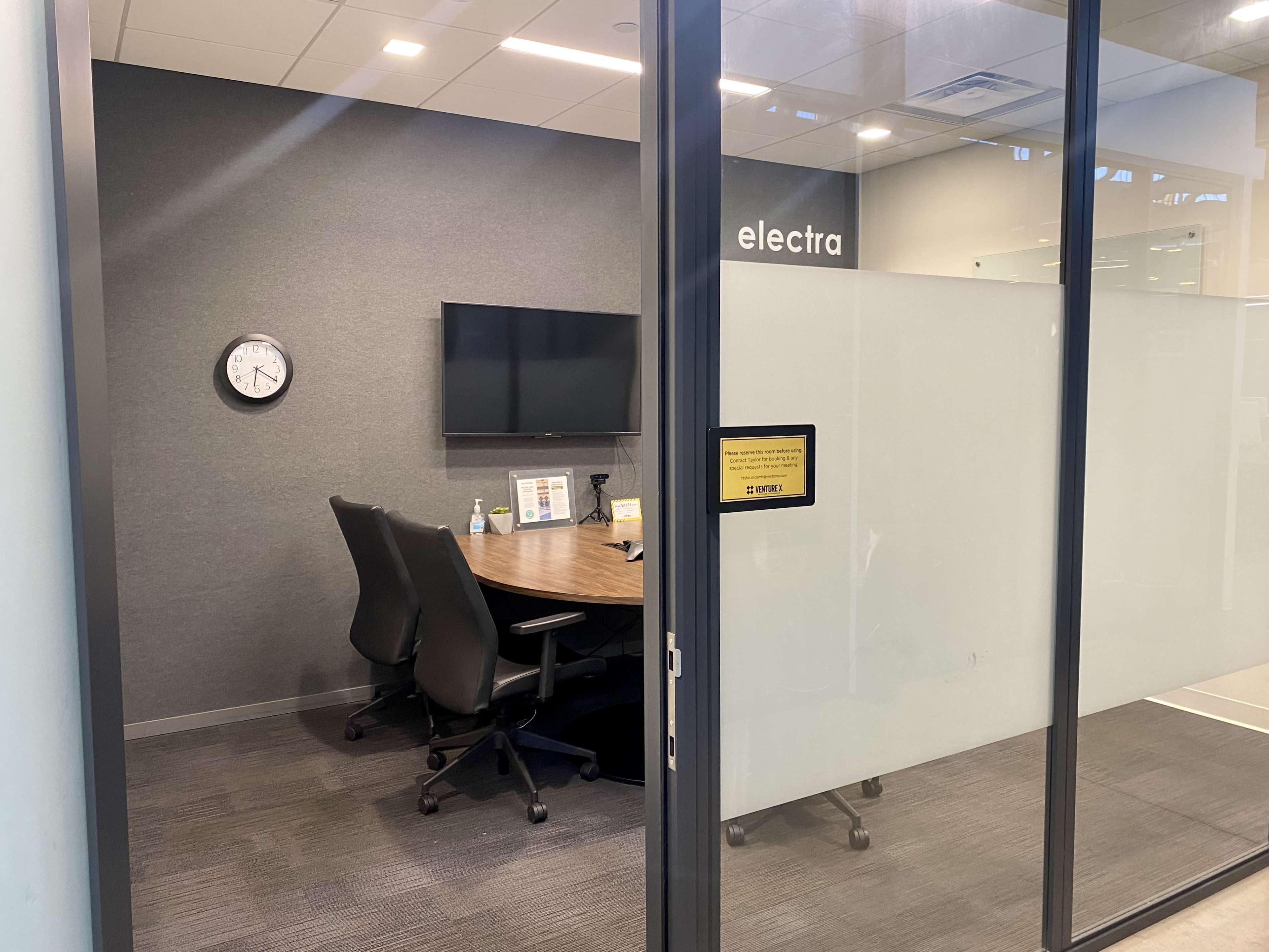 A glass-walled meeting room with a round table, four chairs, a wall-mounted TV, a clock, and a sign on the door.