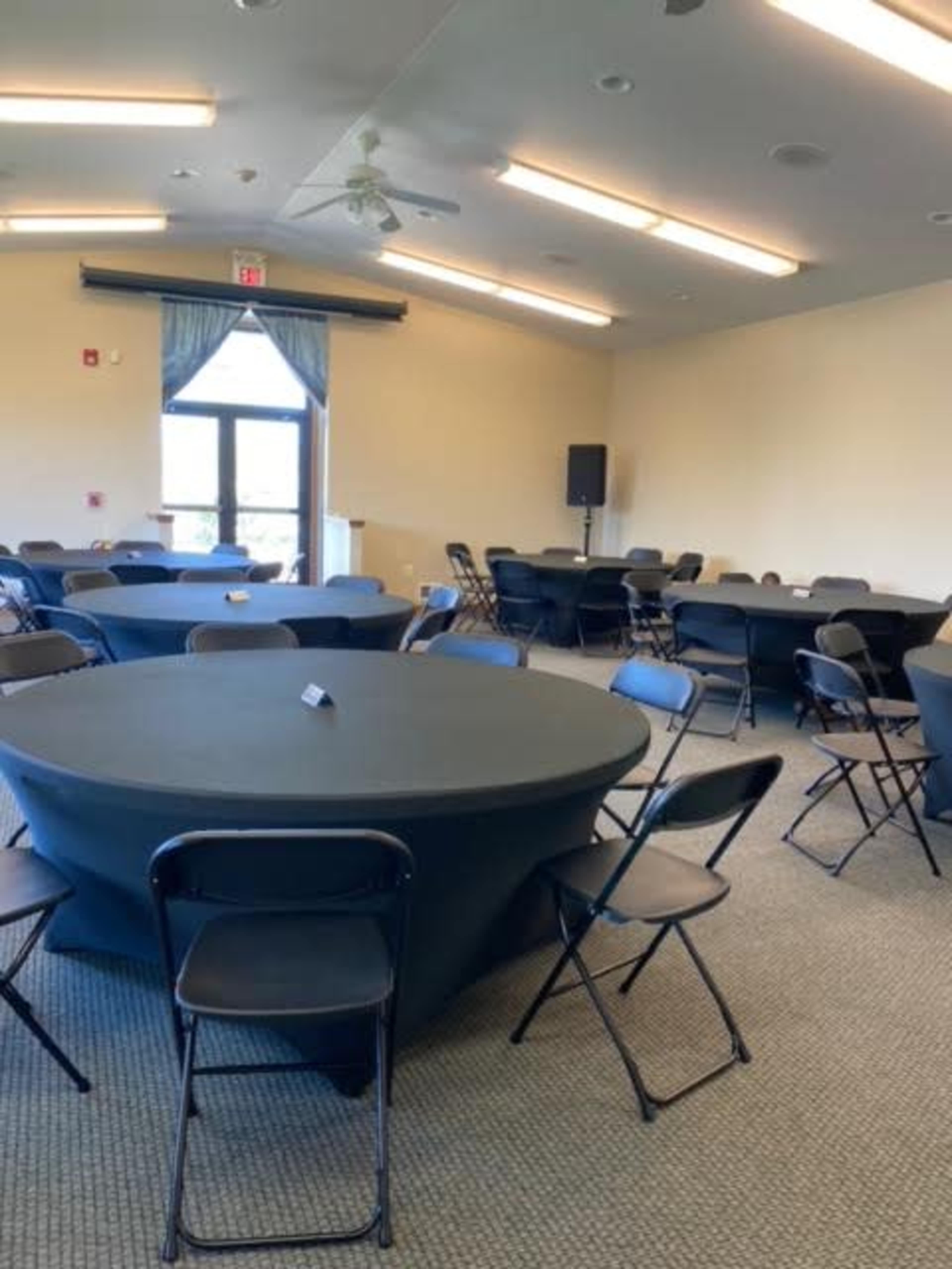 A spacious room with several round tables covered in black cloth and surrounded by folding chairs, illuminated by overhead lights.