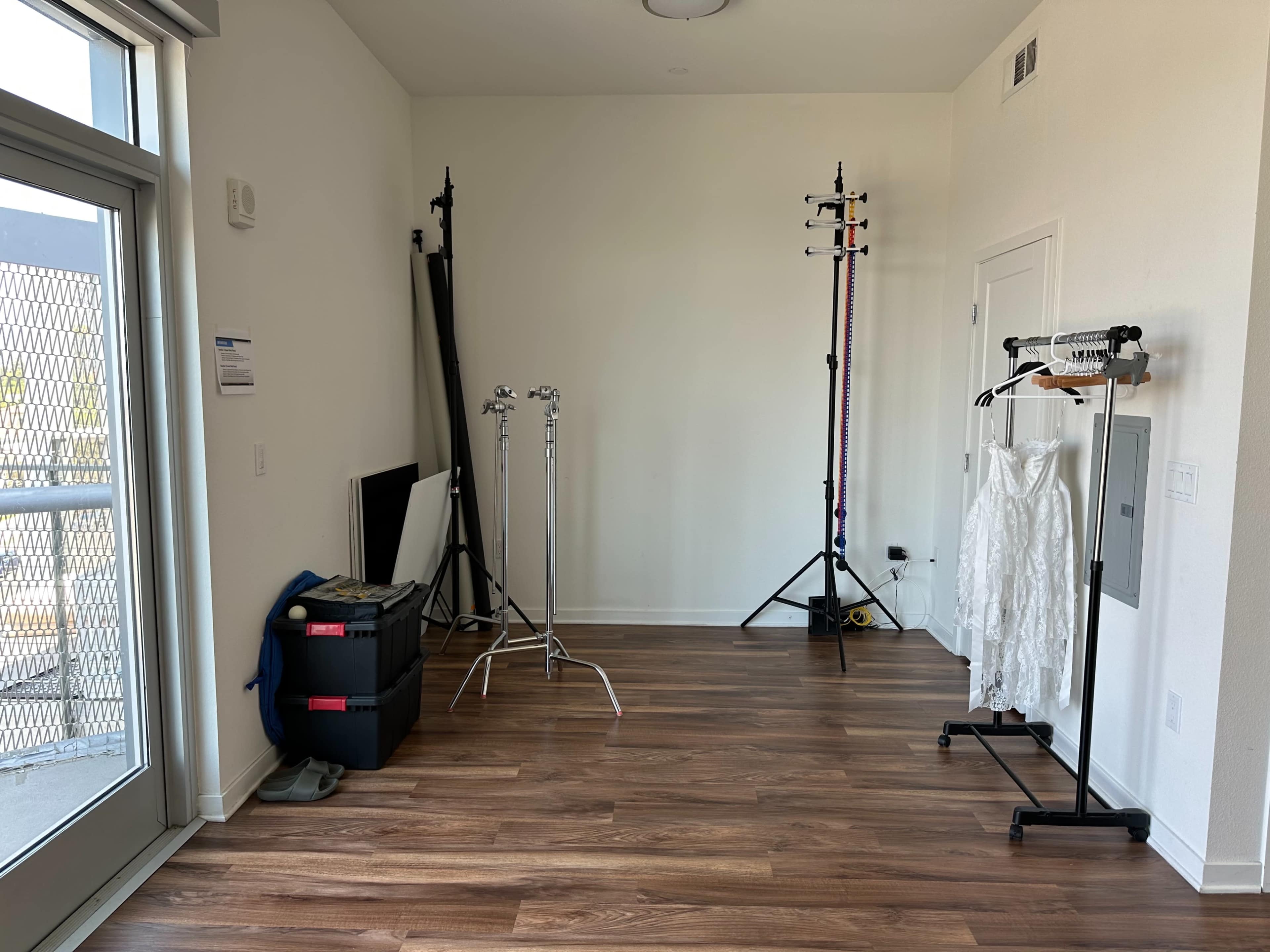The image shows a sparsely furnished room with wooden flooring, featuring clothing racks, lighting stands, and a large window.