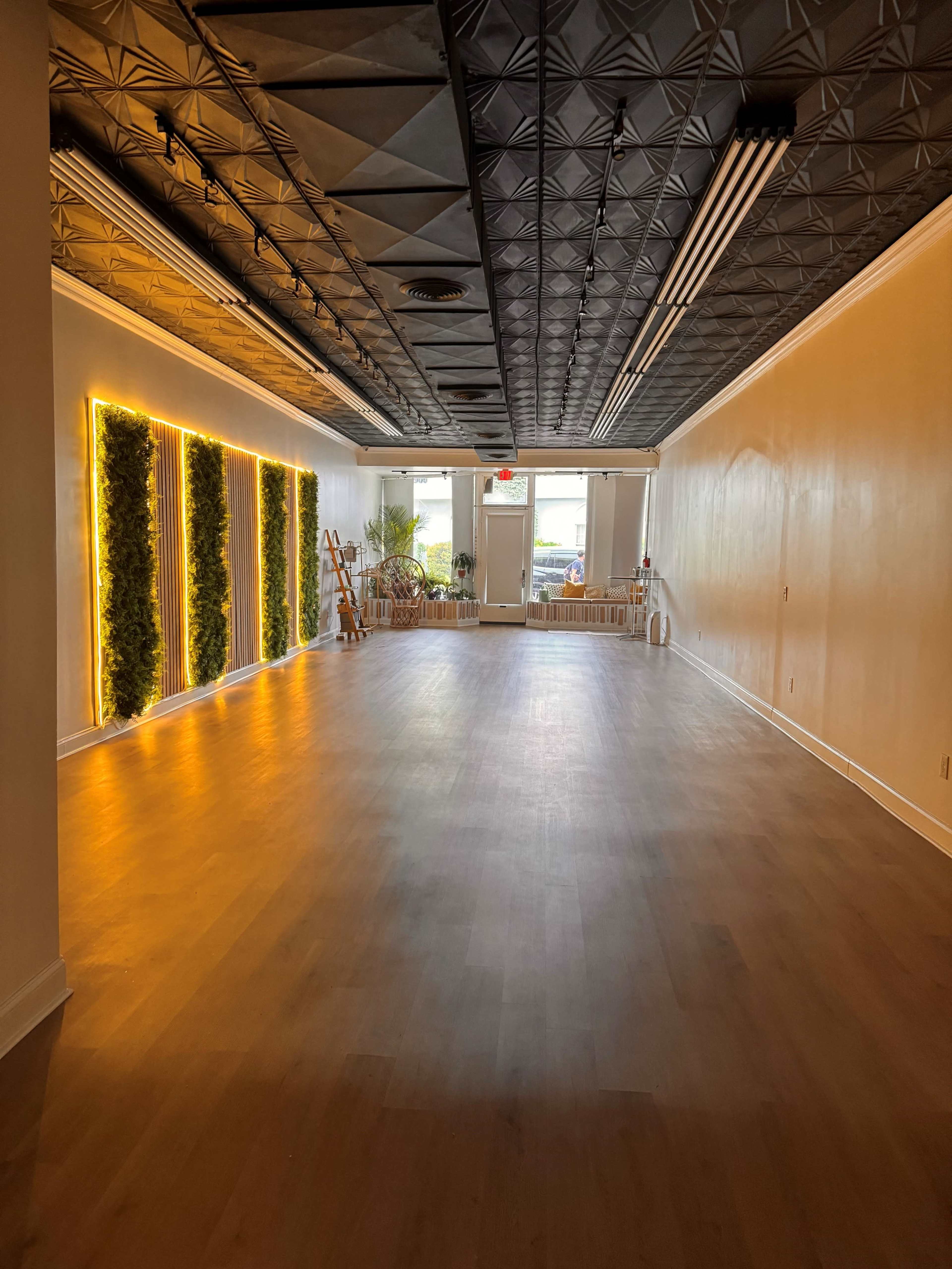 The image shows a spacious, well-lit interior with wooden flooring, featuring decorative wall panels with greenery and a view of large windows at the far end.