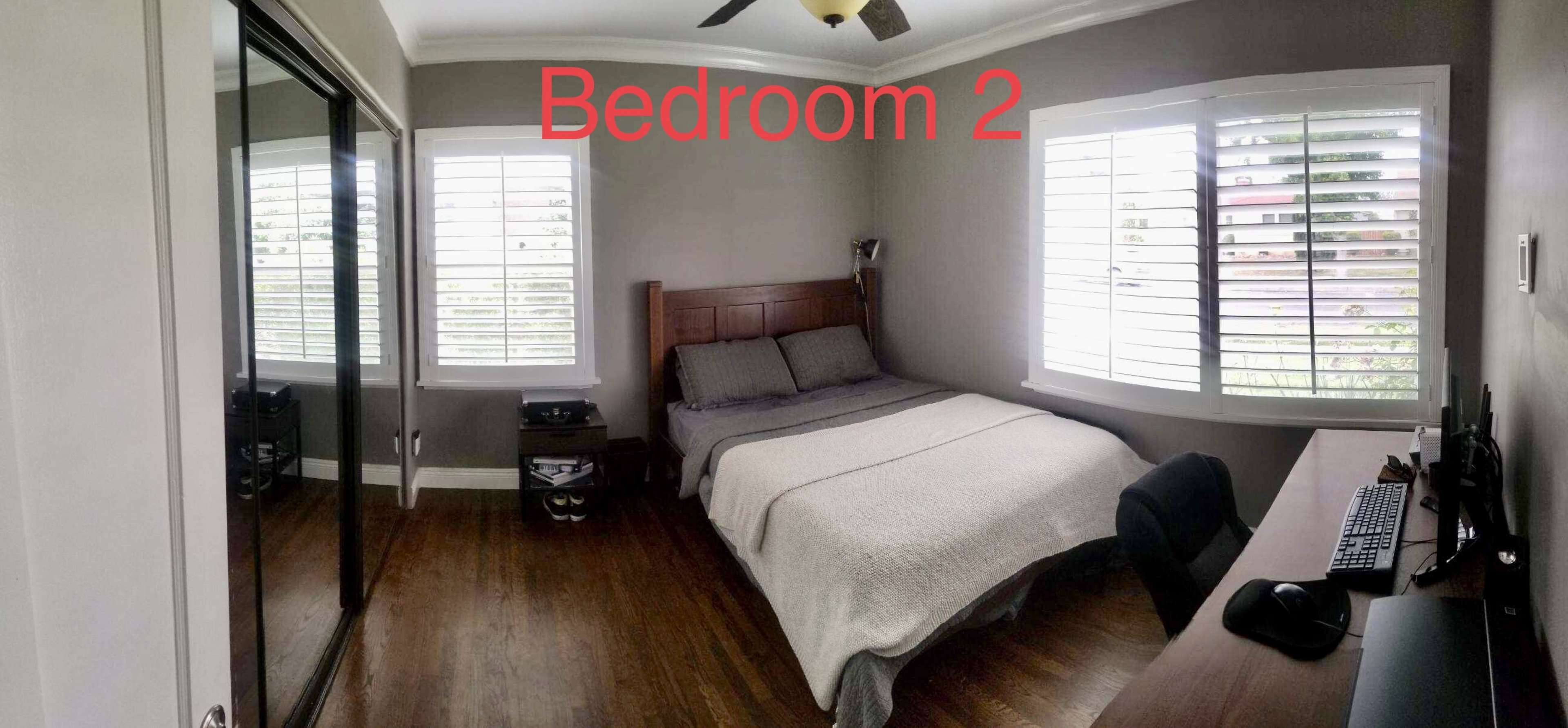 A modern bedroom features a bed with a gray blanket, a large mirror on one wall, and a desk with a computer setup.