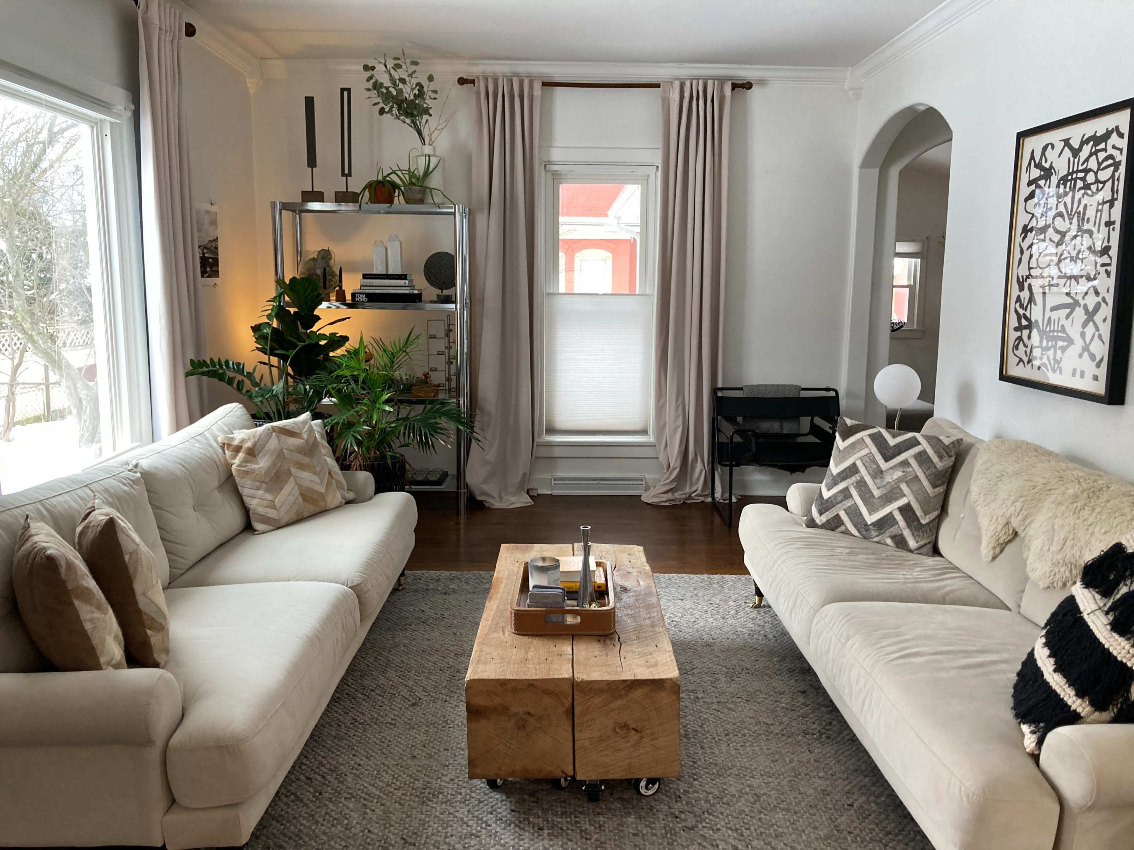 A modern living room featuring two white sofas, a wooden coffee table on wheels, a large area rug, and a window with sheer curtains.