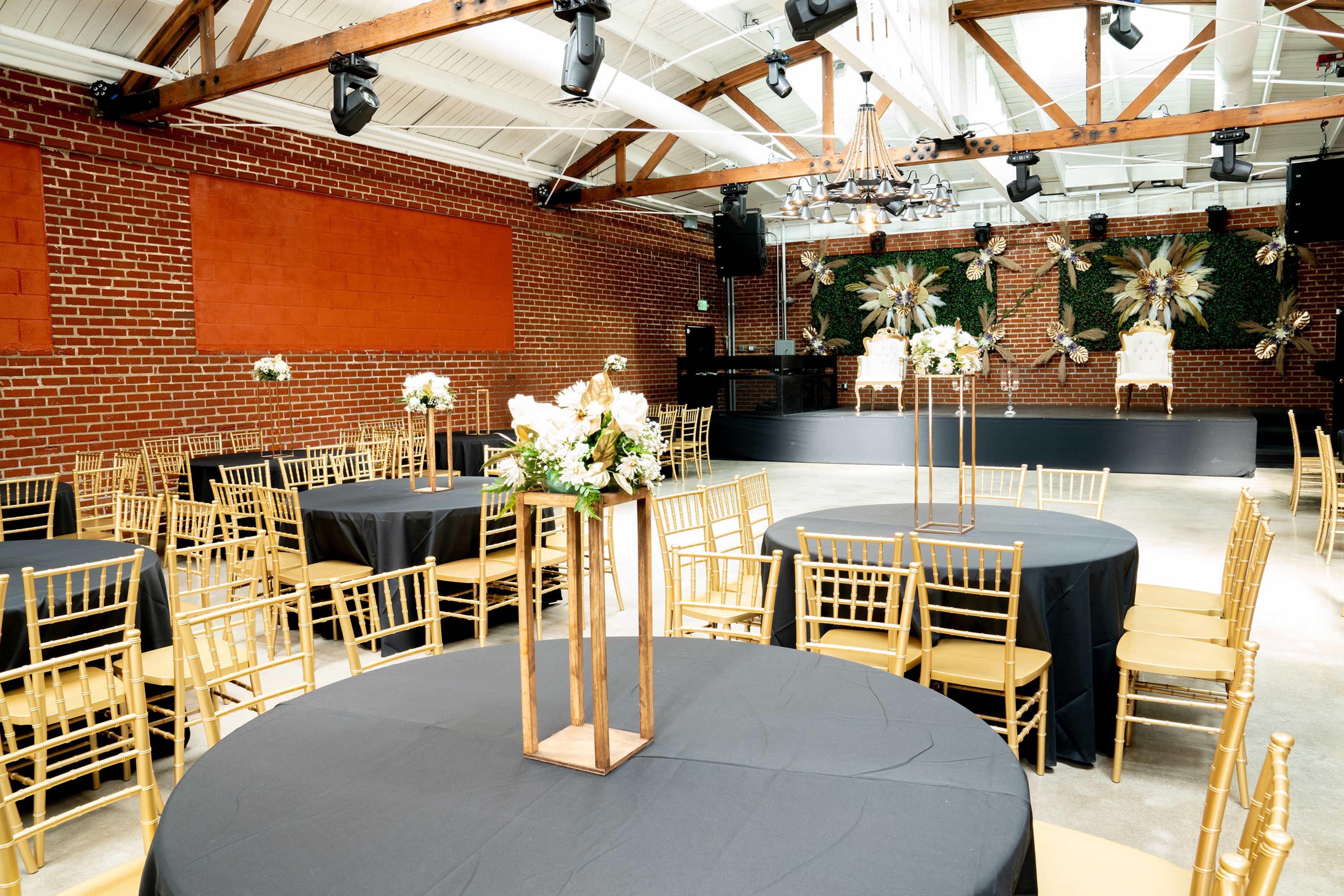 The image shows a spacious event venue with round tables covered in black tablecloths, decorated with floral centerpieces, and a stage at the front featuring elegant chairs and wall decor.