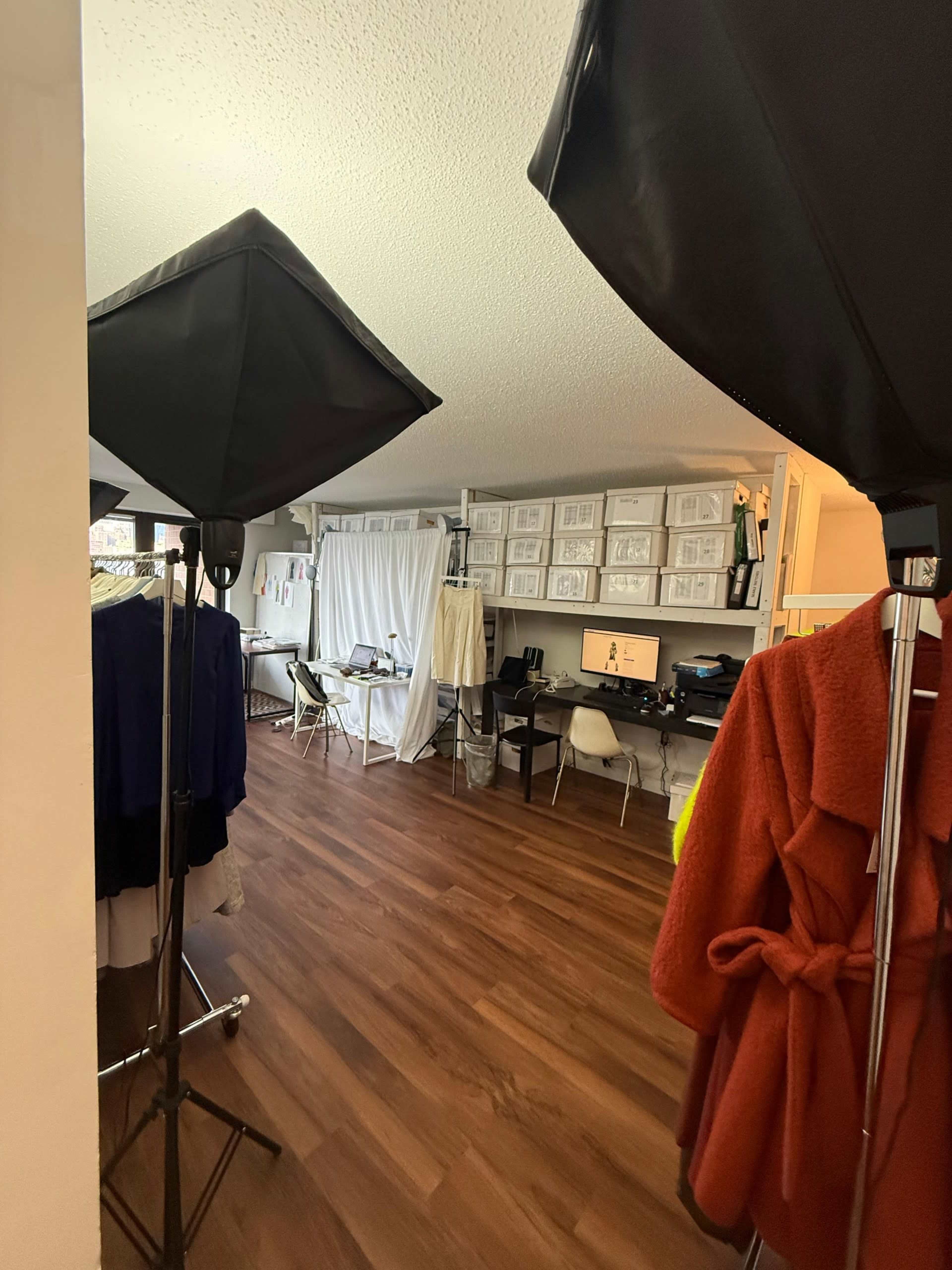 The image shows a spacious room set up for a photoshoot, featuring lighting equipment, clothing racks, and a desk with a computer.