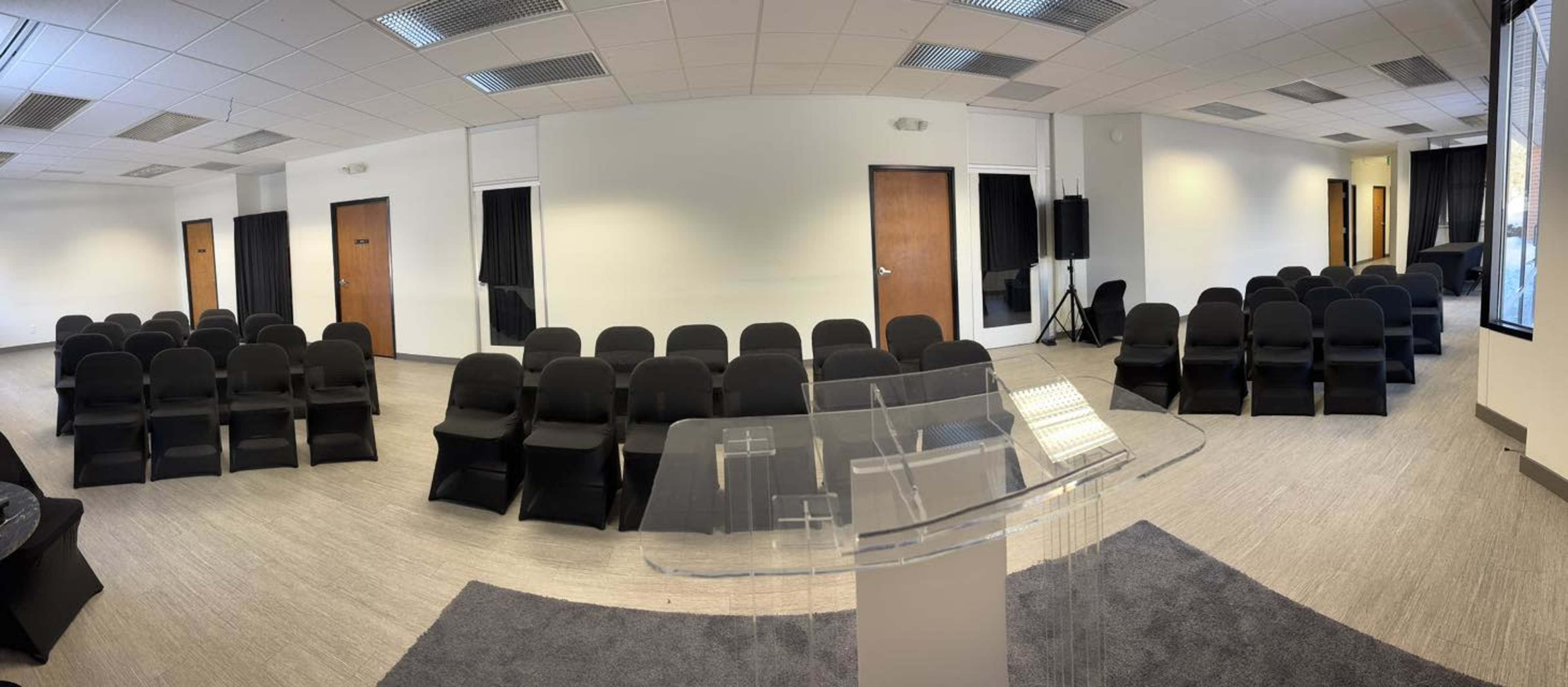 The image shows a spacious room arranged with rows of black chairs facing a clear podium in the foreground.