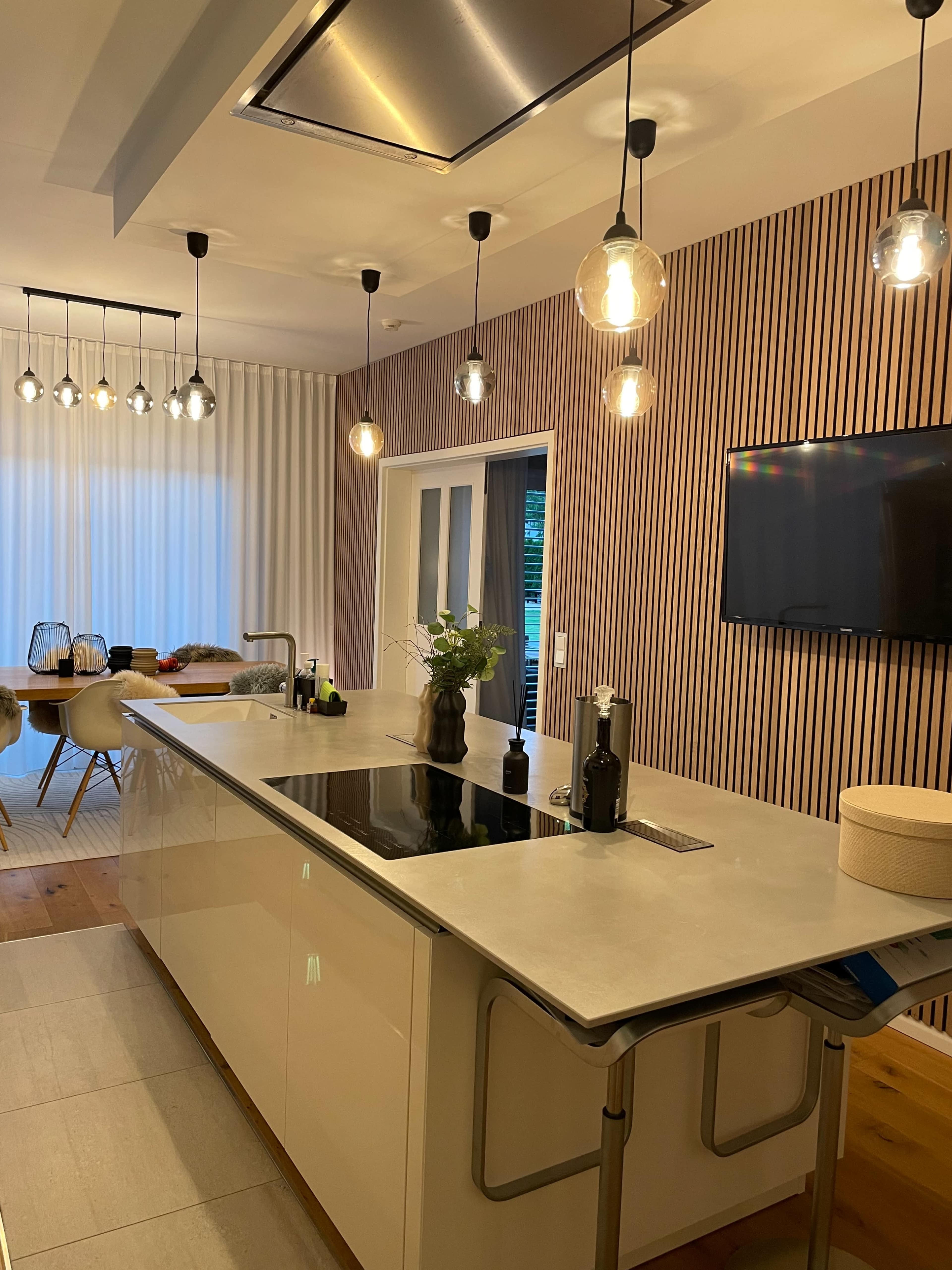 A modern kitchen with a central island surrounded by pendant lights, featuring a sleek countertop and a wall-mounted television.