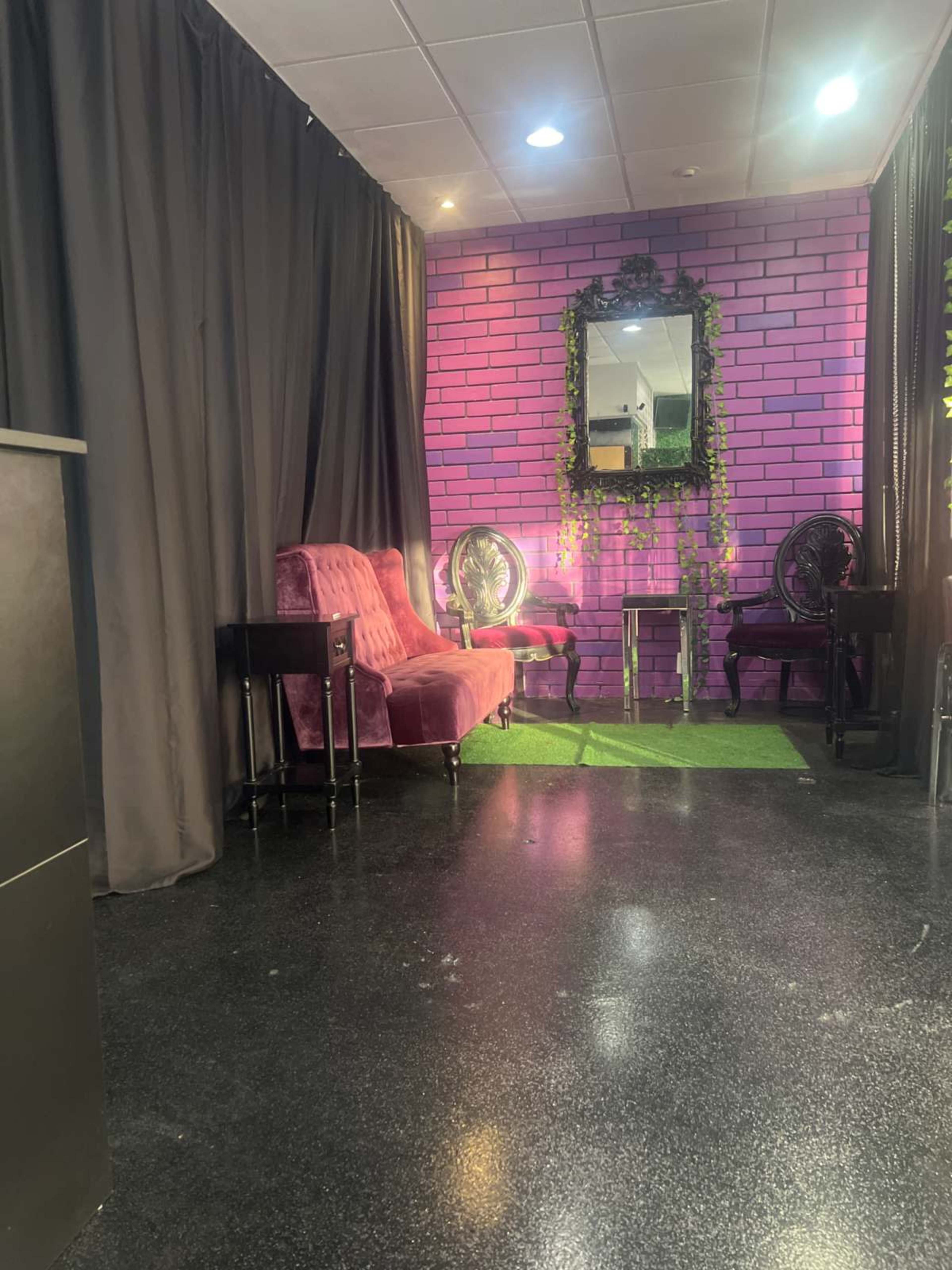 The image shows a small interior space featuring a pink-brick wall, velvet seating, a mirror, and decorative plants, with dark curtains framing the area.