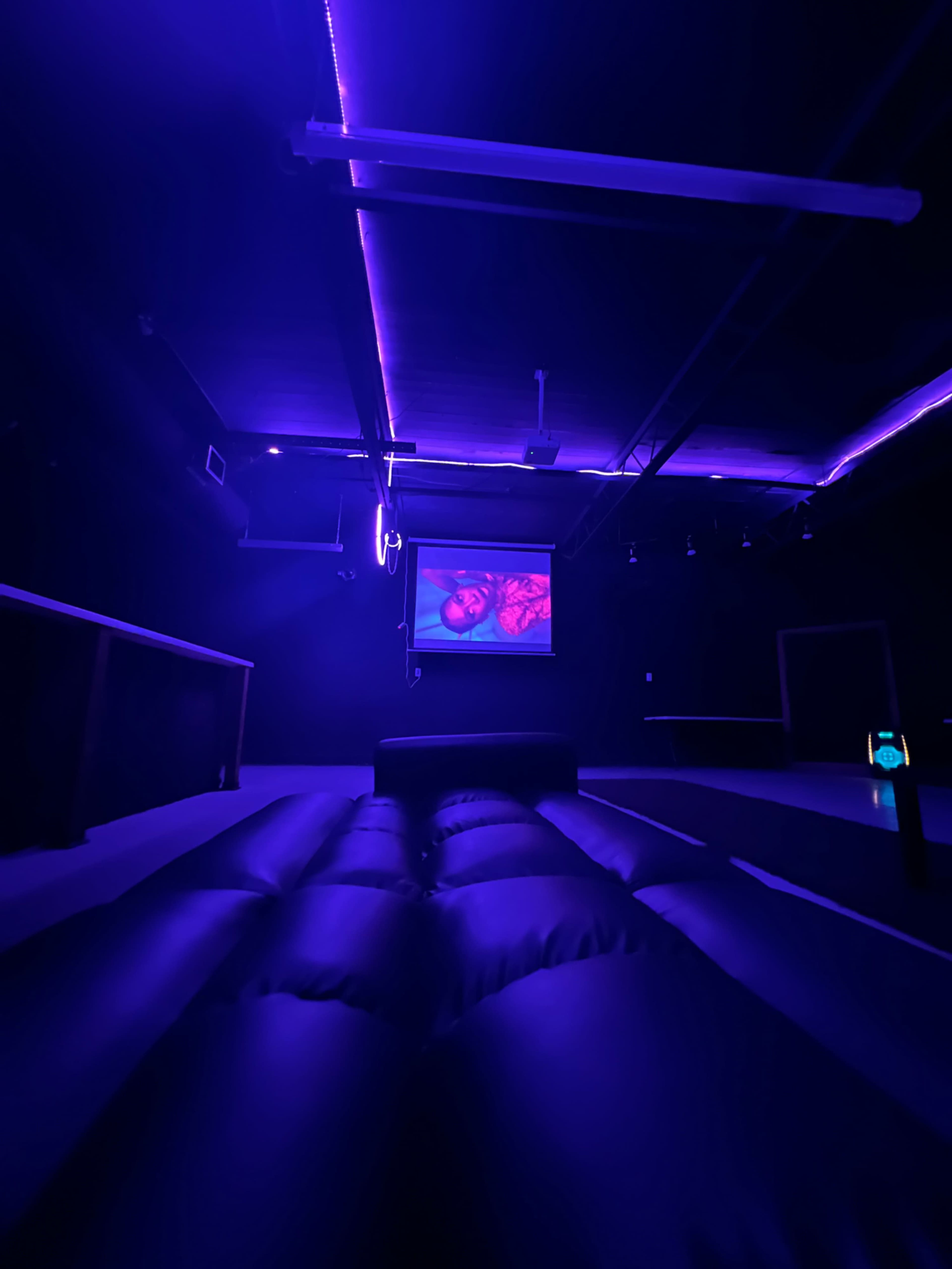 A dimly lit room features a large screen displaying an image, with purple lighting illuminating a soft, inflated seating area.