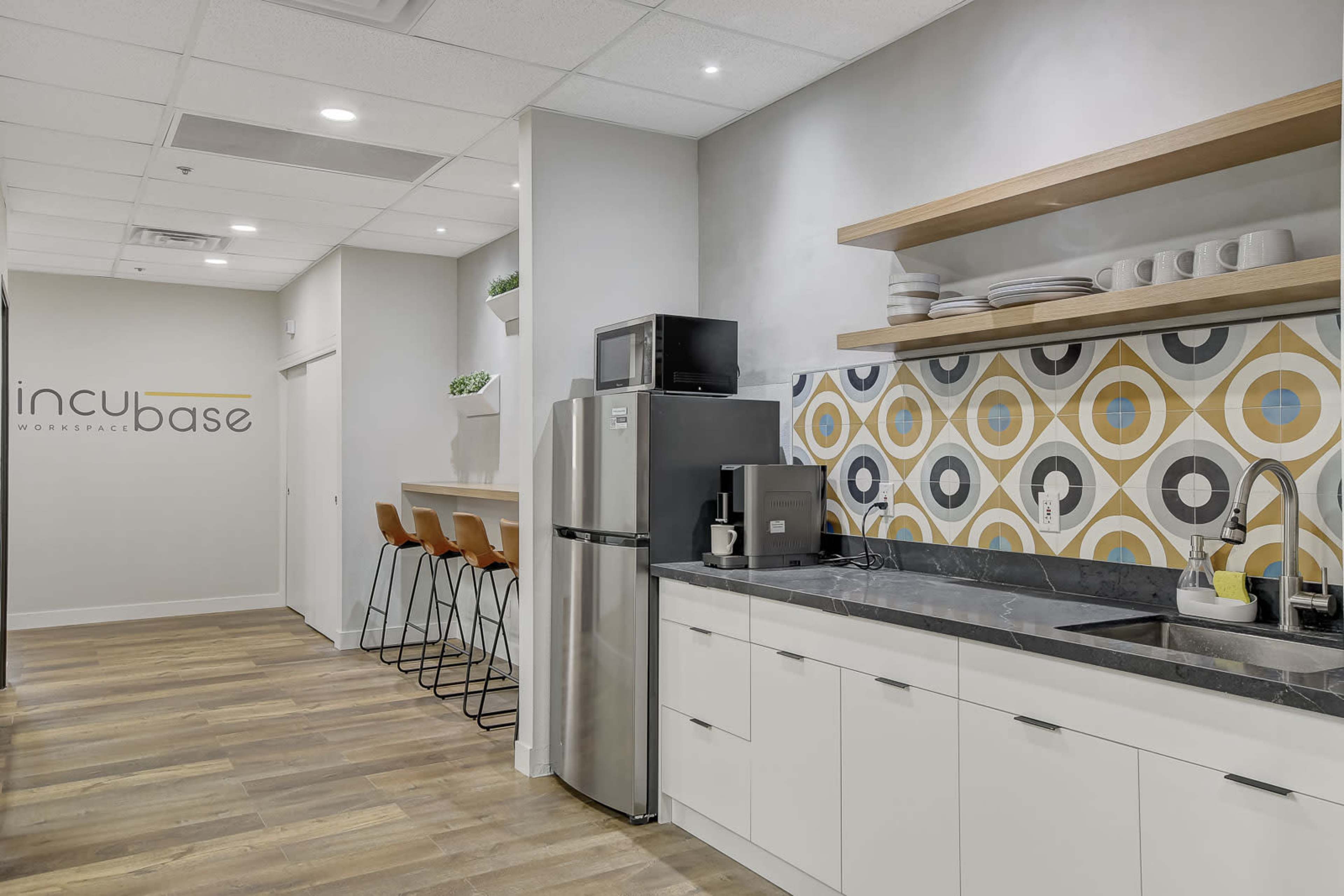 A modern kitchen area within a workspace, featuring a refrigerator, sink, and colorful geometric-patterned backsplash, alongside a counter with bar stools.