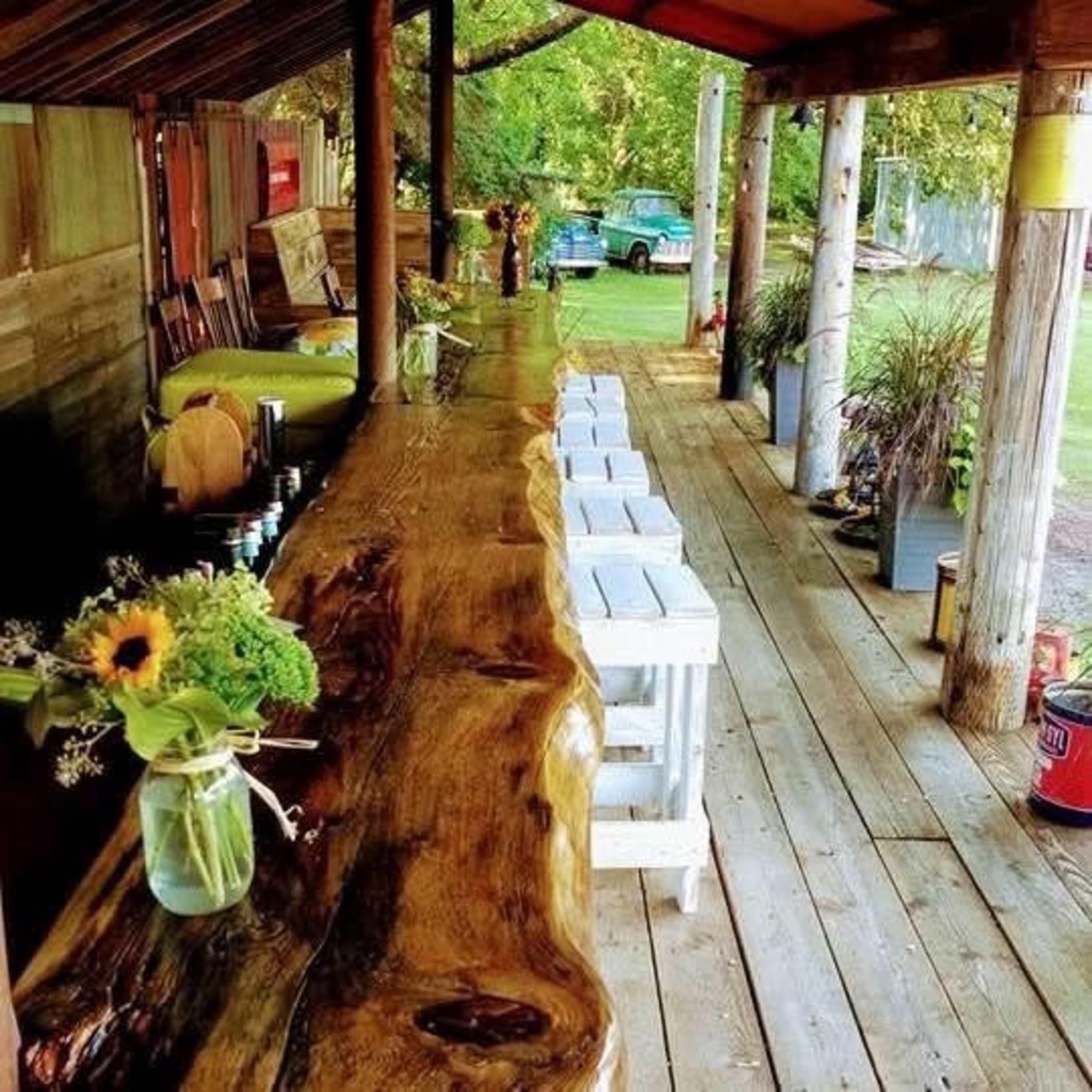 The image shows a wooden bar area with a natural edge countertop, surrounded by white stools and filled with greenery, under a rustic roof in a backyard setting.