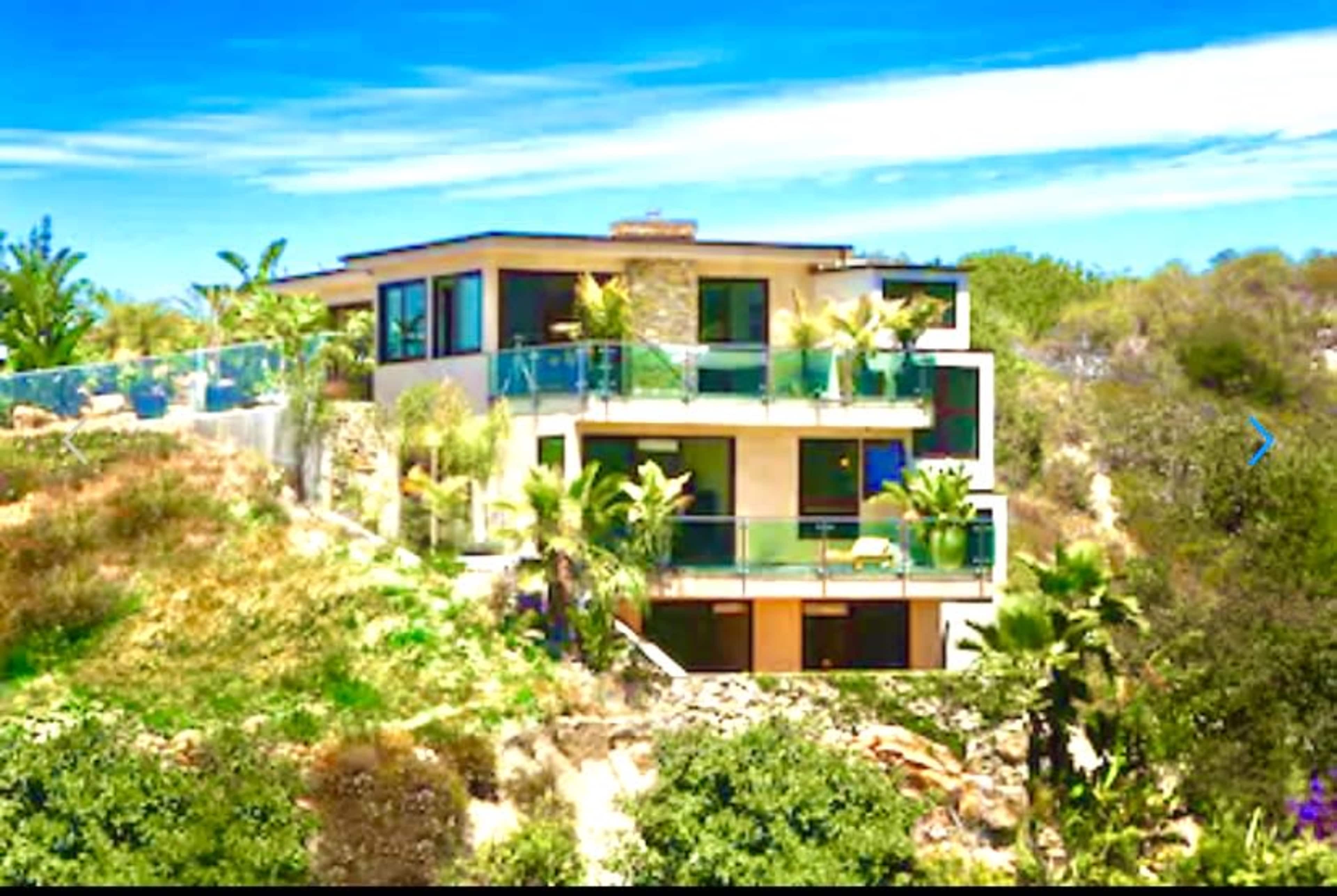 A modern three-story house is situated on a hillside, surrounded by lush greenery and palm trees, with large glass balconies.