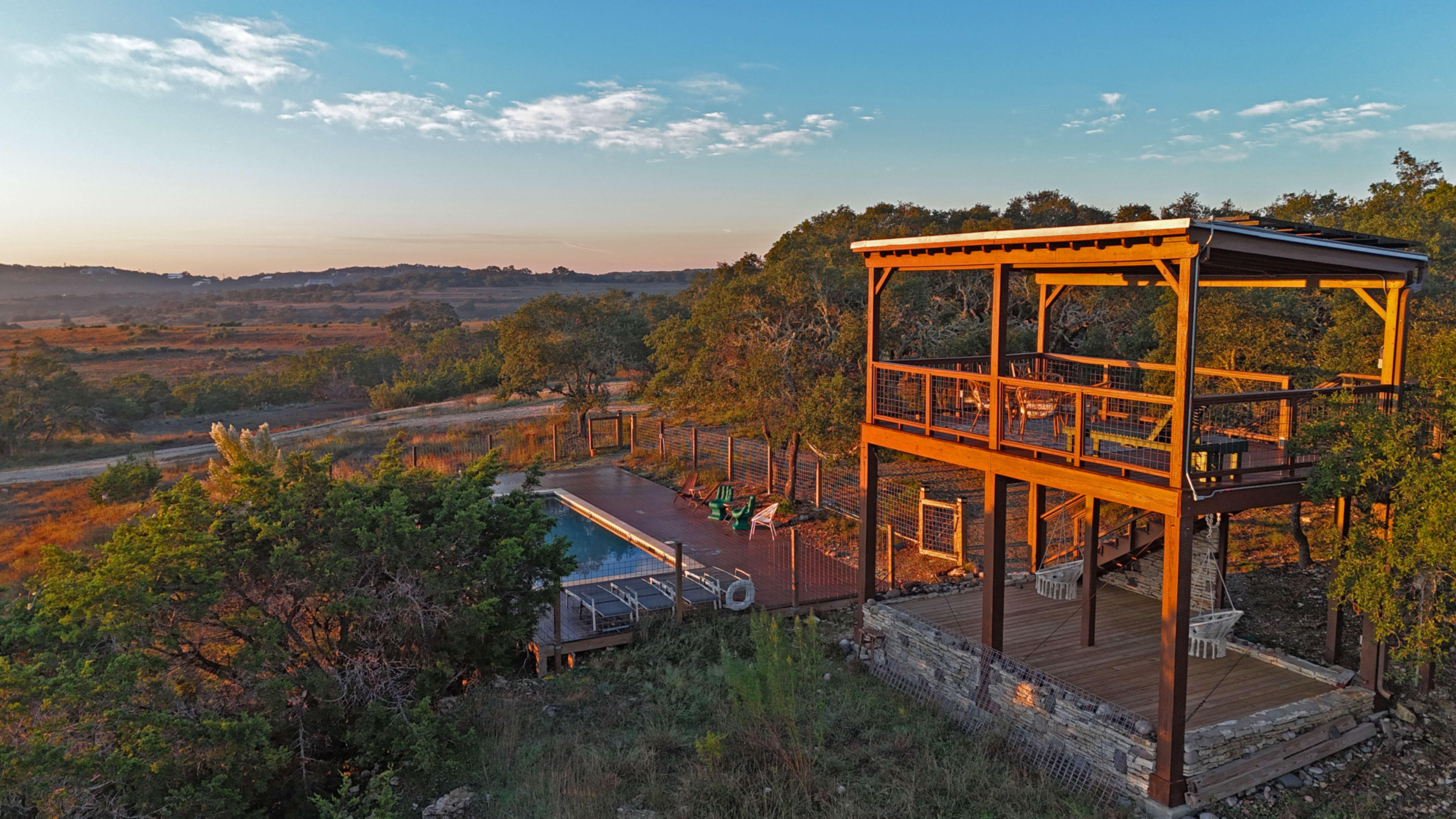 A wooden deck with seating overlooks a pool and a landscape of rolling hills at sunset.