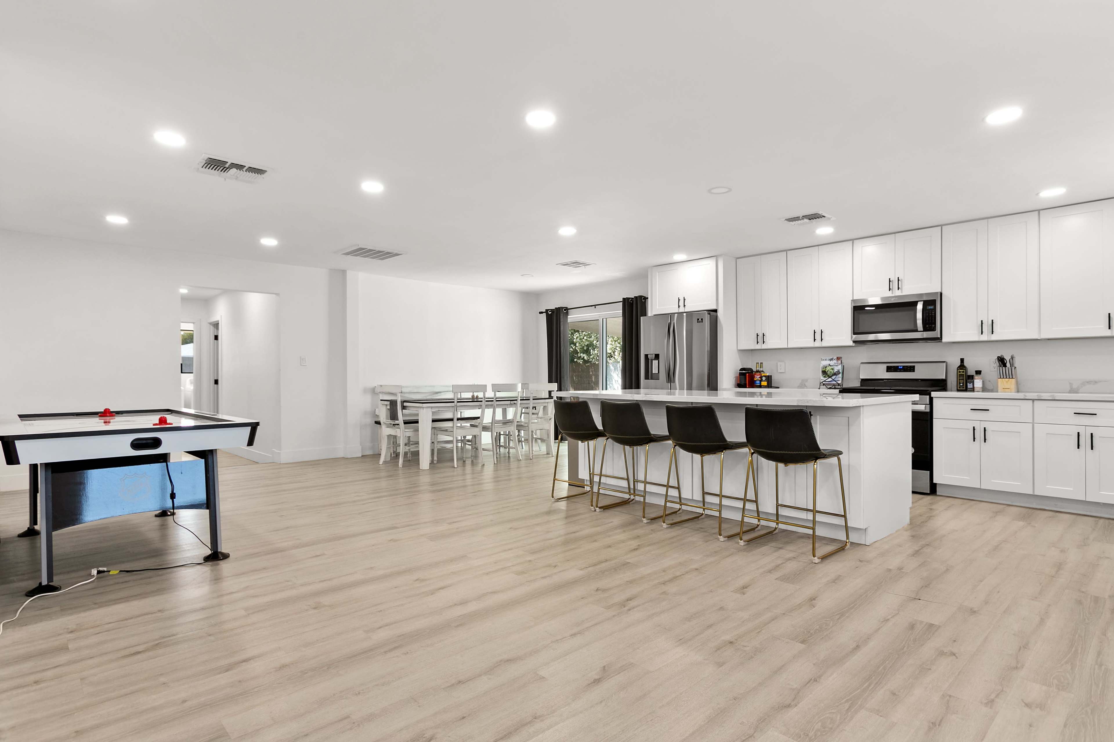 The image shows a modern open-concept kitchen and living area featuring a large island with bar stools, a dining table, and a game table in a well-lit space.