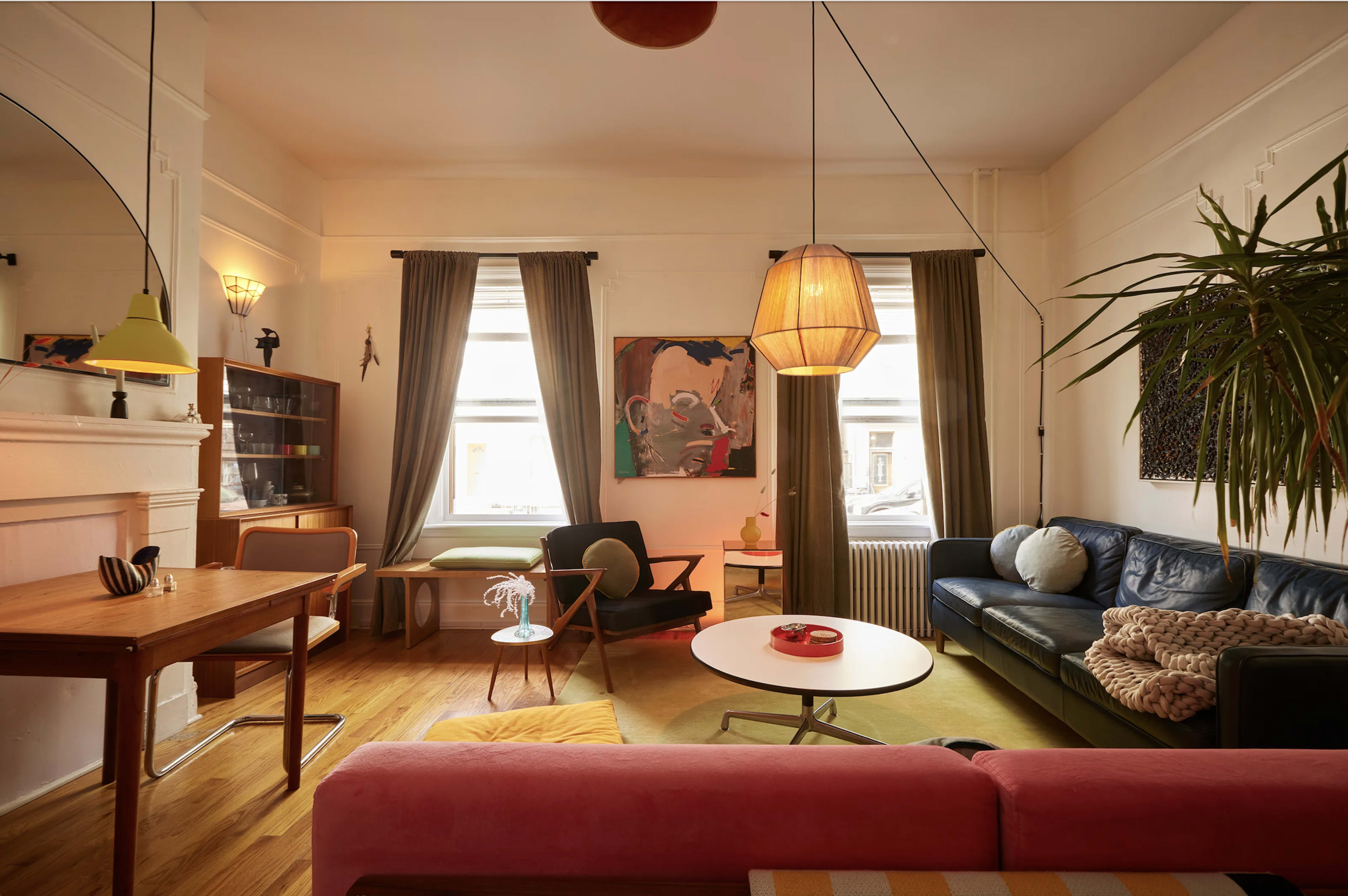 The image shows a stylish living room with a mix of modern and vintage furniture, featuring a blue leather sofa, a pink couch, and a wooden dining table under warm lighting.