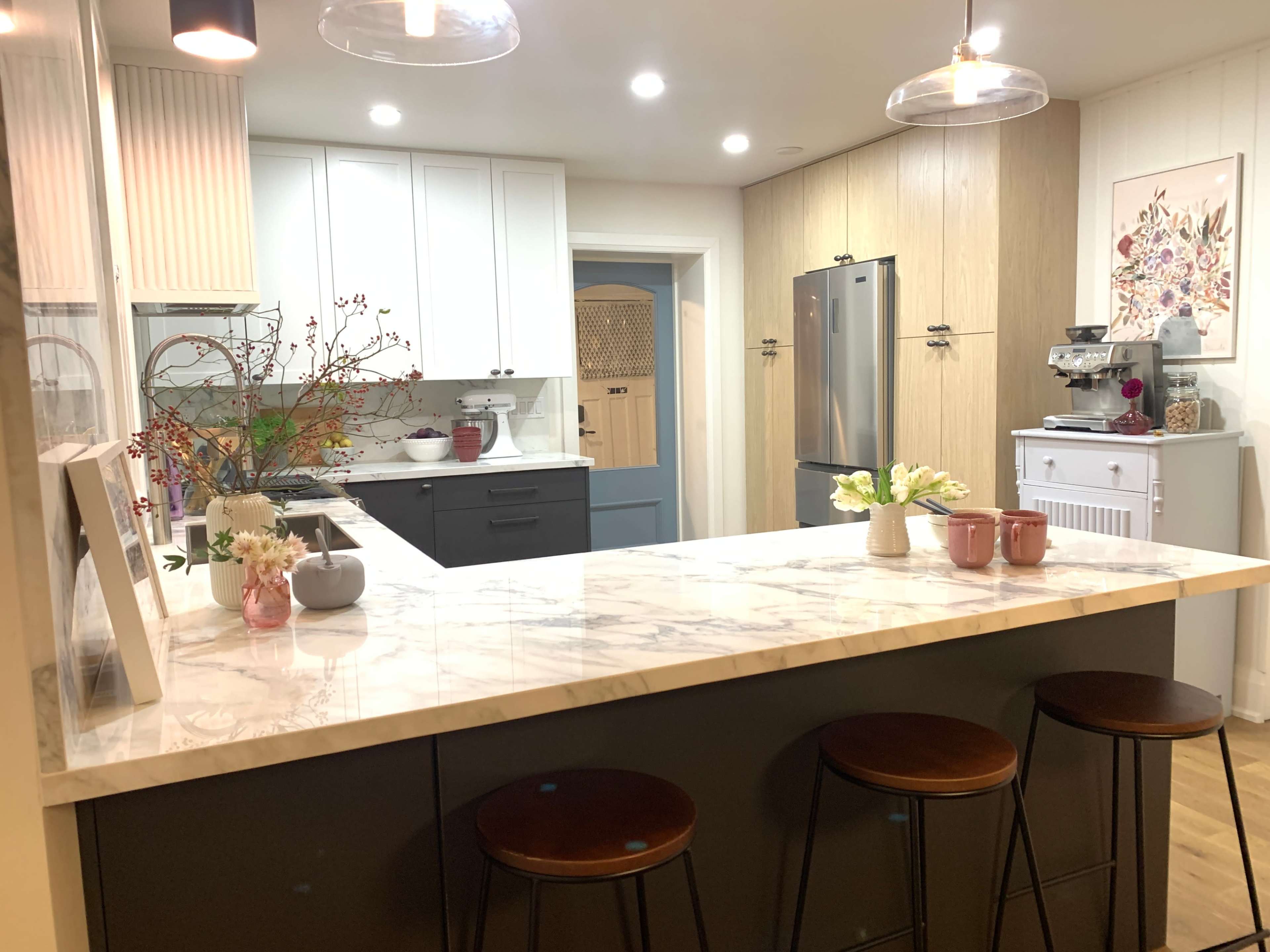 A modern kitchen features a marble countertop island with three wooden stools, stainless steel appliances, and a mix of light cabinetry.