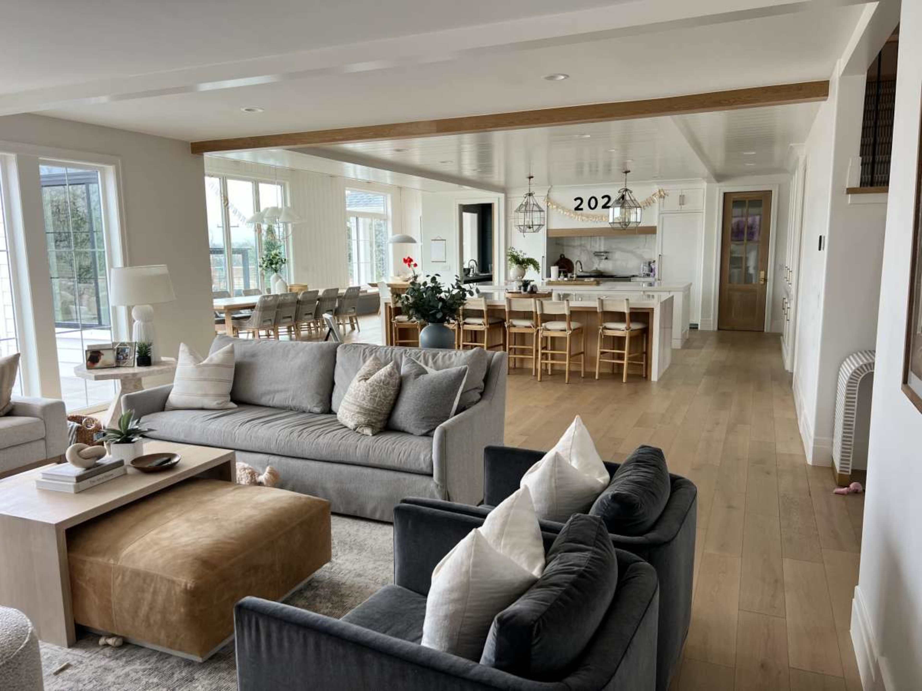 The image shows a spacious, modern living area with two gray sofas, a wooden coffee table, and an open kitchen in the background featuring light-colored cabinetry and several bar stools.