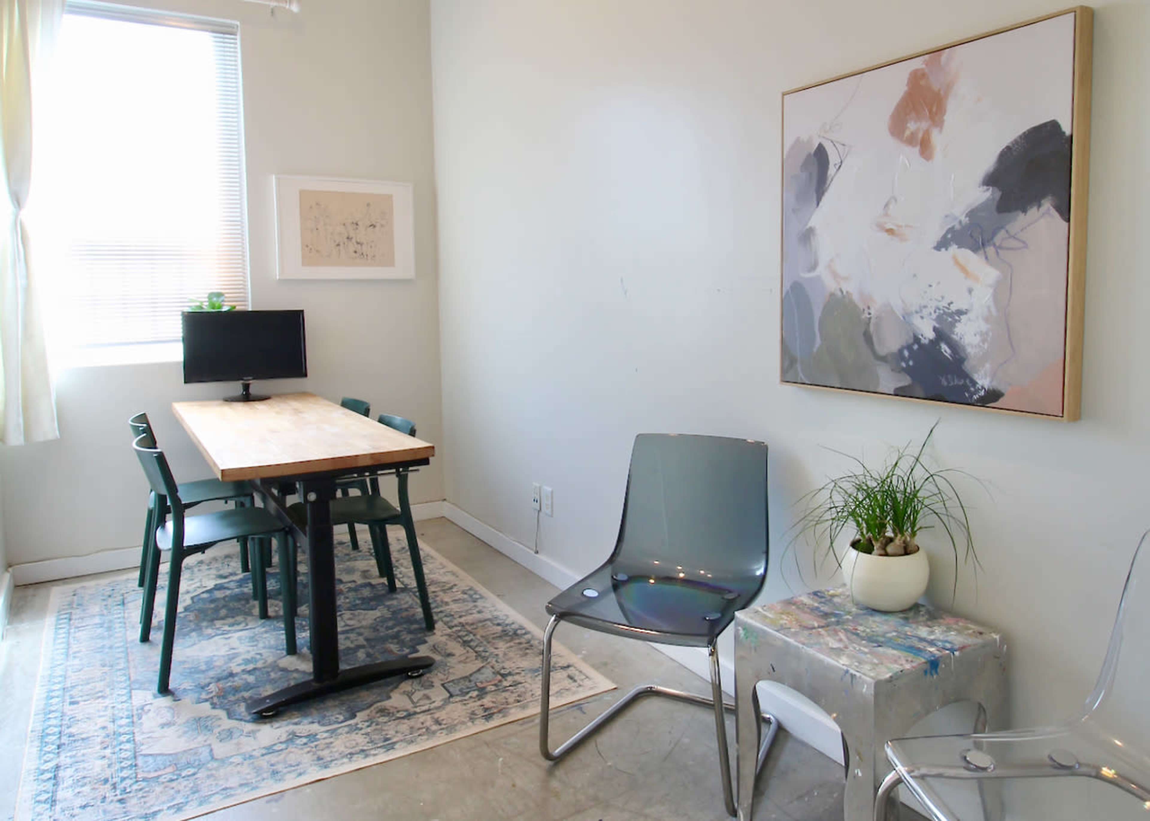 The image shows a minimalistic workspace with a wooden table, green chairs, a computer monitor, a decorative plant, and an abstract painting on the wall.