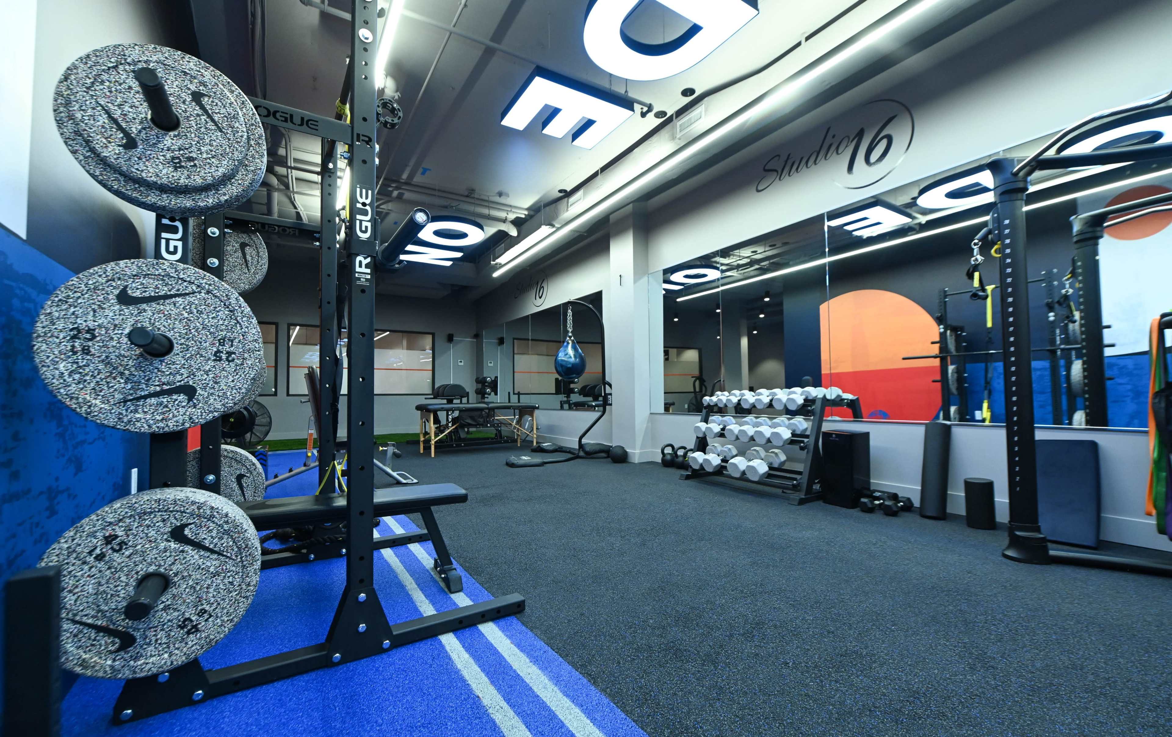 The image shows a modern fitness studio equipped with weightlifting racks, dumbbells, and a punching bag.