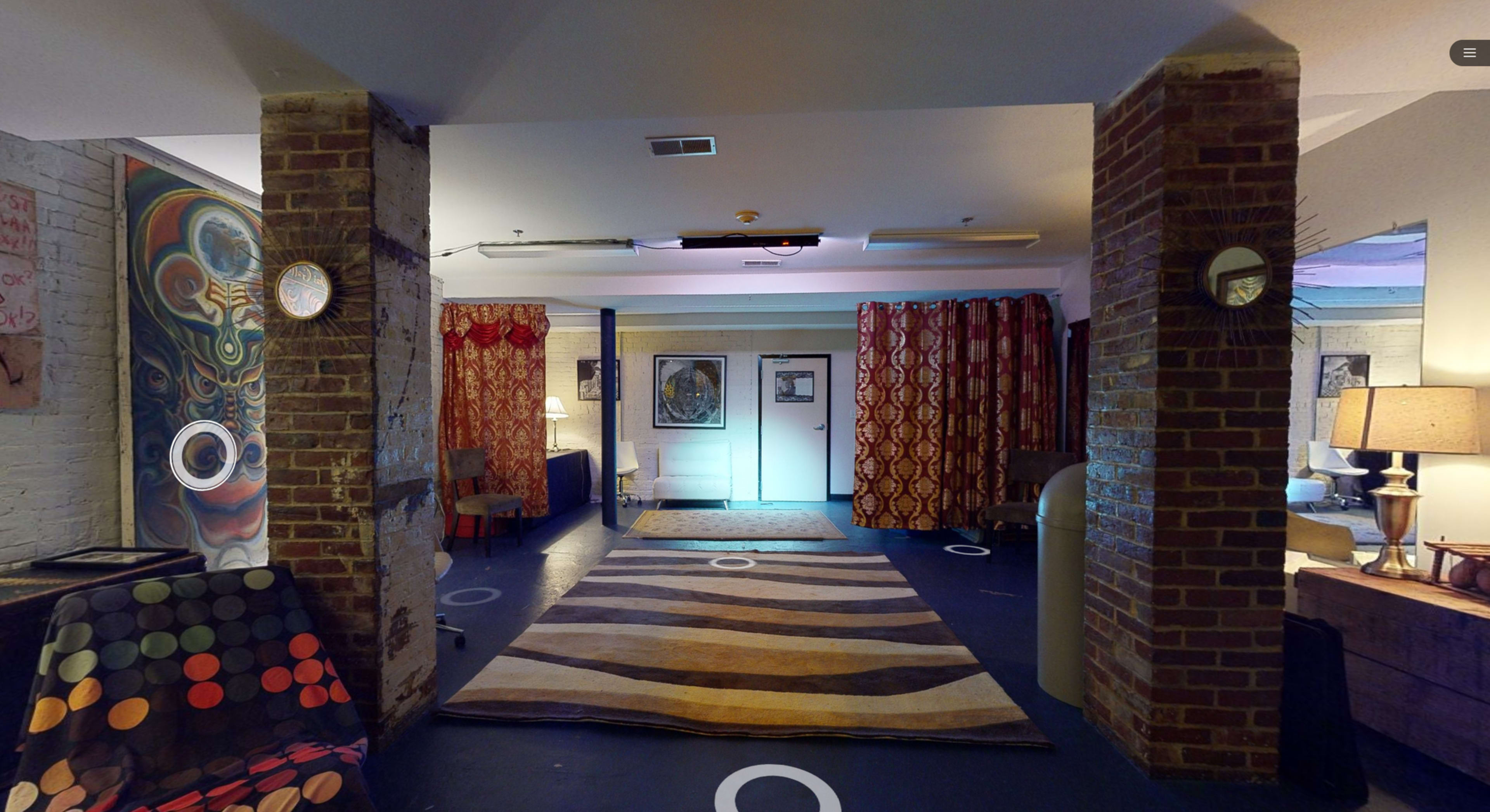 The image shows a spacious, lit room with patterned curtains, a striped rug, and artwork on the walls, featuring two brick columns.