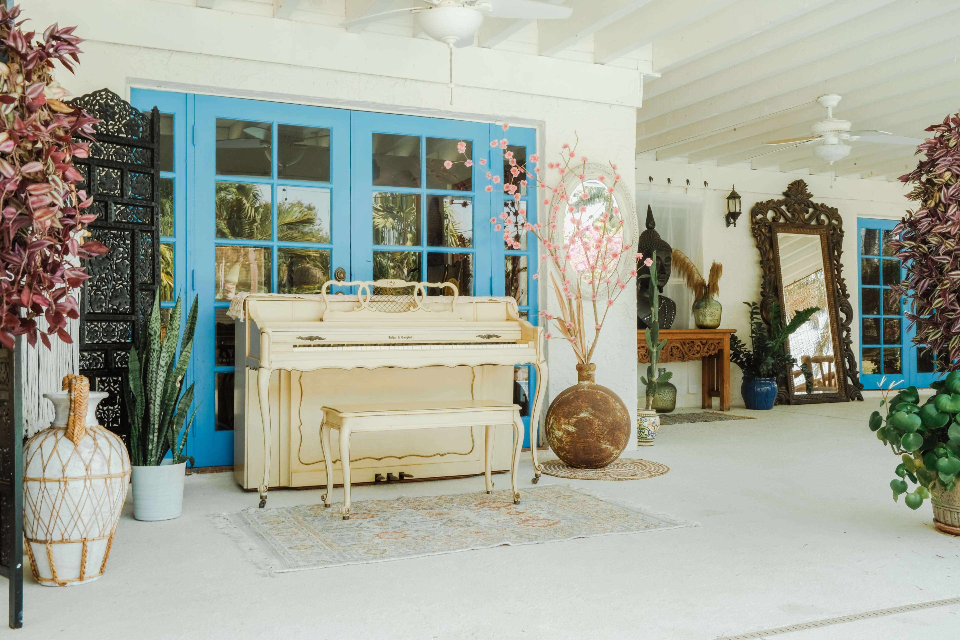 The image features a light-colored piano and bench positioned in a bright, airy space with blue French doors and various decorative plants.