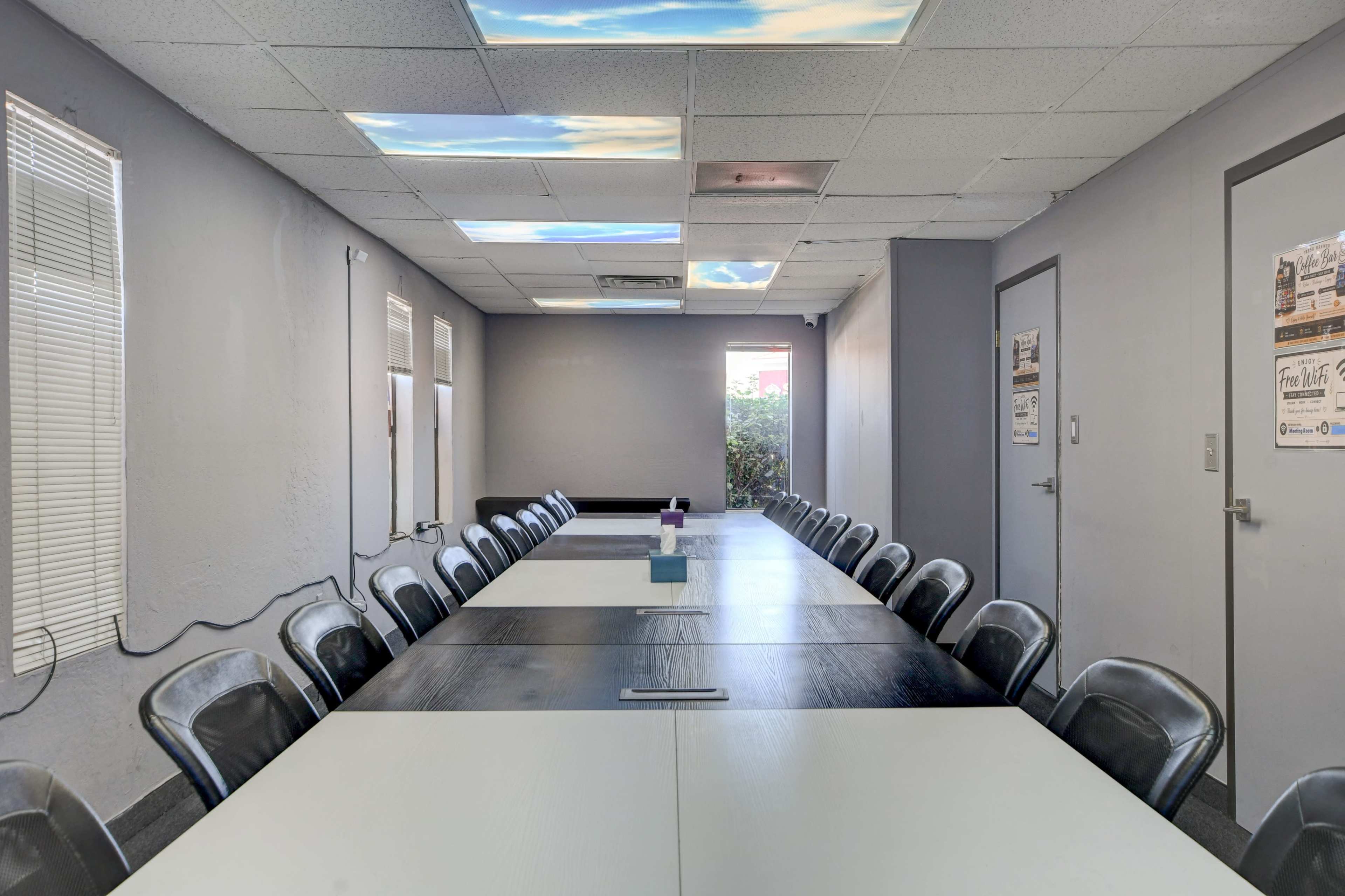 A large, rectangular meeting room features a long table surrounded by black chairs, illuminated by artificial lighting and a mural of a blue sky on the ceiling.
