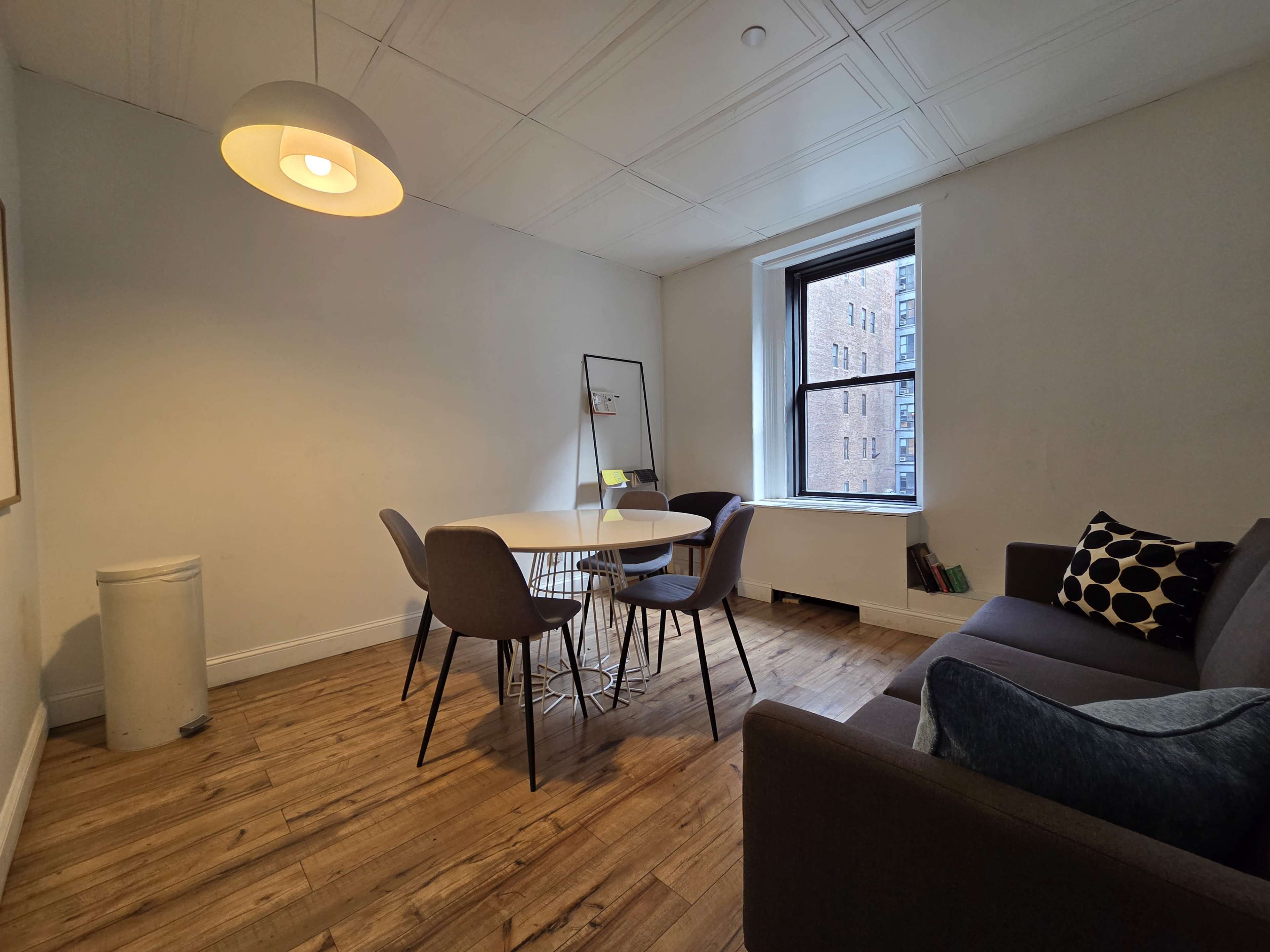 A small, well-lit room features a round dining table with four chairs, a sofa, a trash can, and a large window overlooking a brick building.