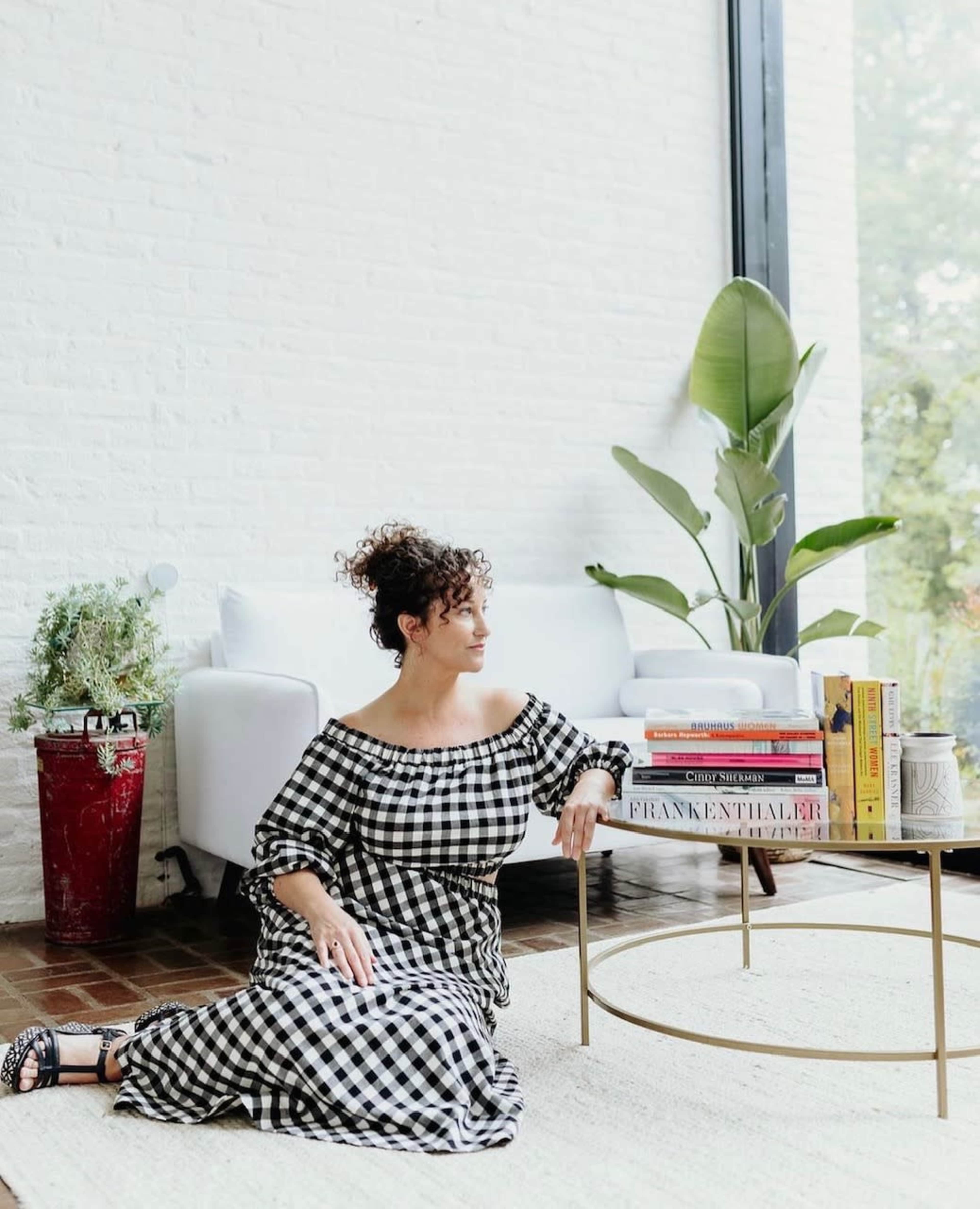 A woman in a checked dress sits on the floor beside a coffee table stacked with books, in a room featuring a large window and a potted plant.