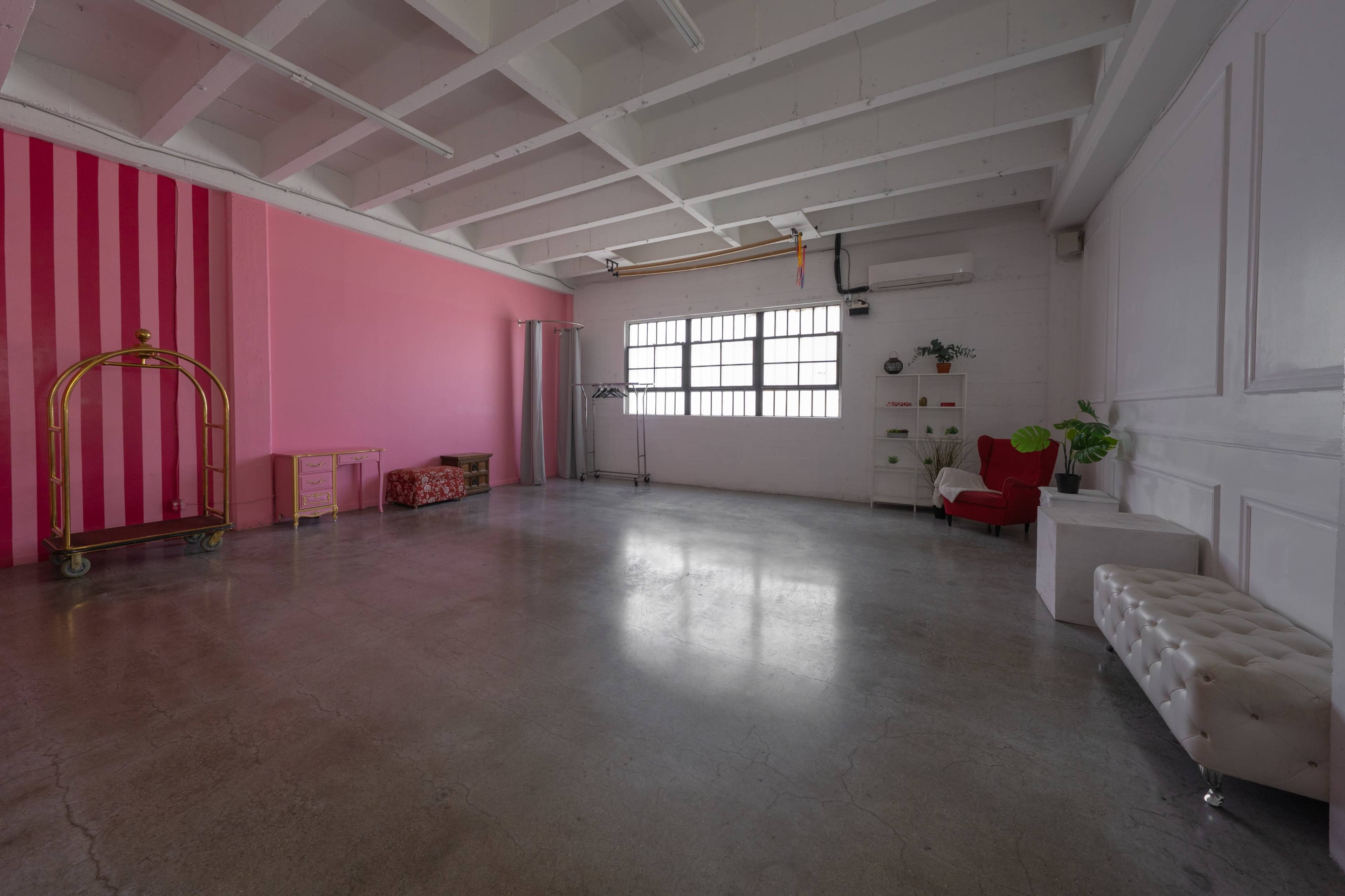 The image shows an empty room with concrete flooring, a pink striped wall, a window with metal bars, and various furniture pieces including a dressing rack, a red chair, and a white tufted bench.