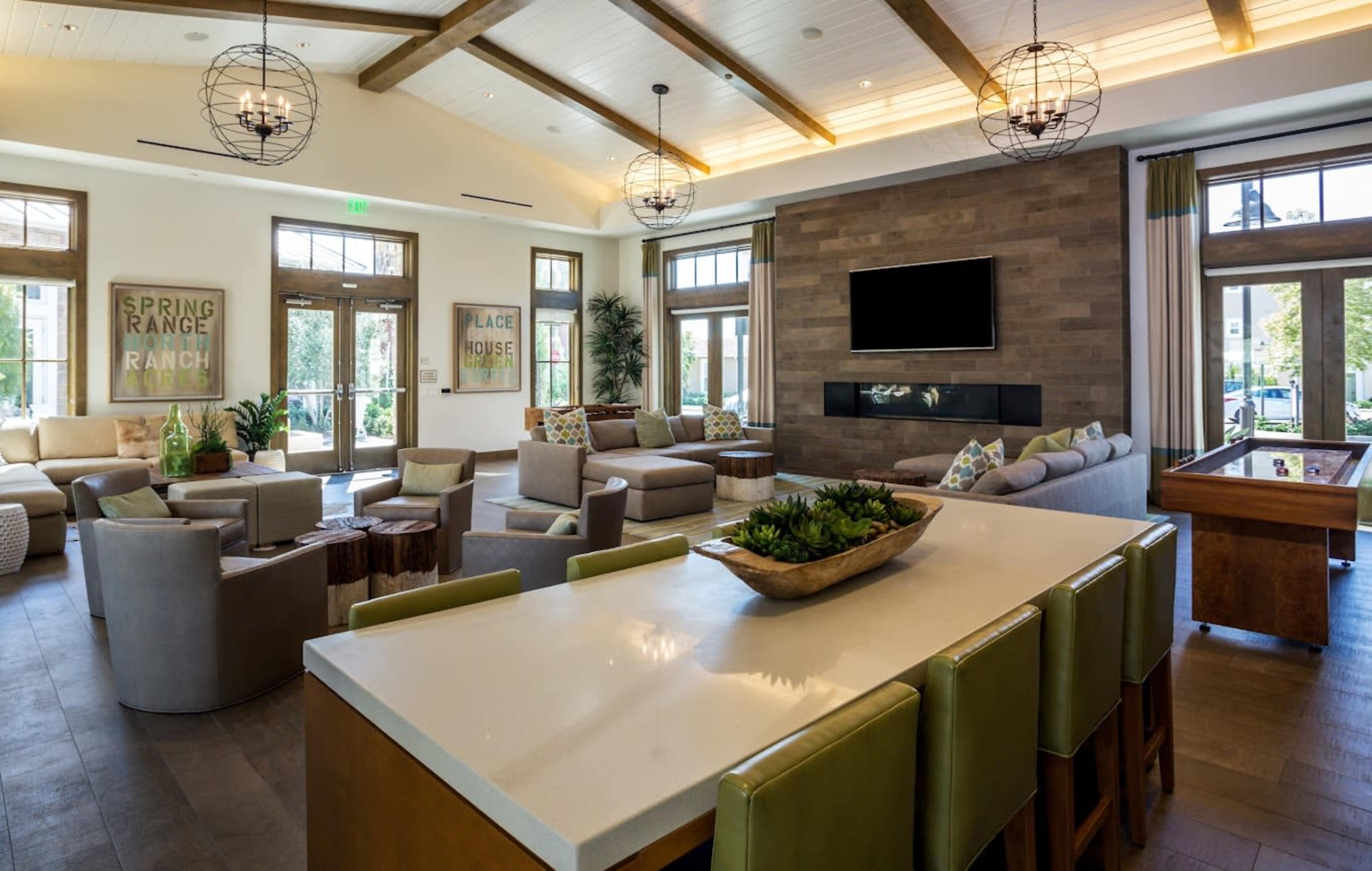 The image shows a modern lounge area with various seating arrangements, a large wall-mounted television, and a kitchen island with bar seating.