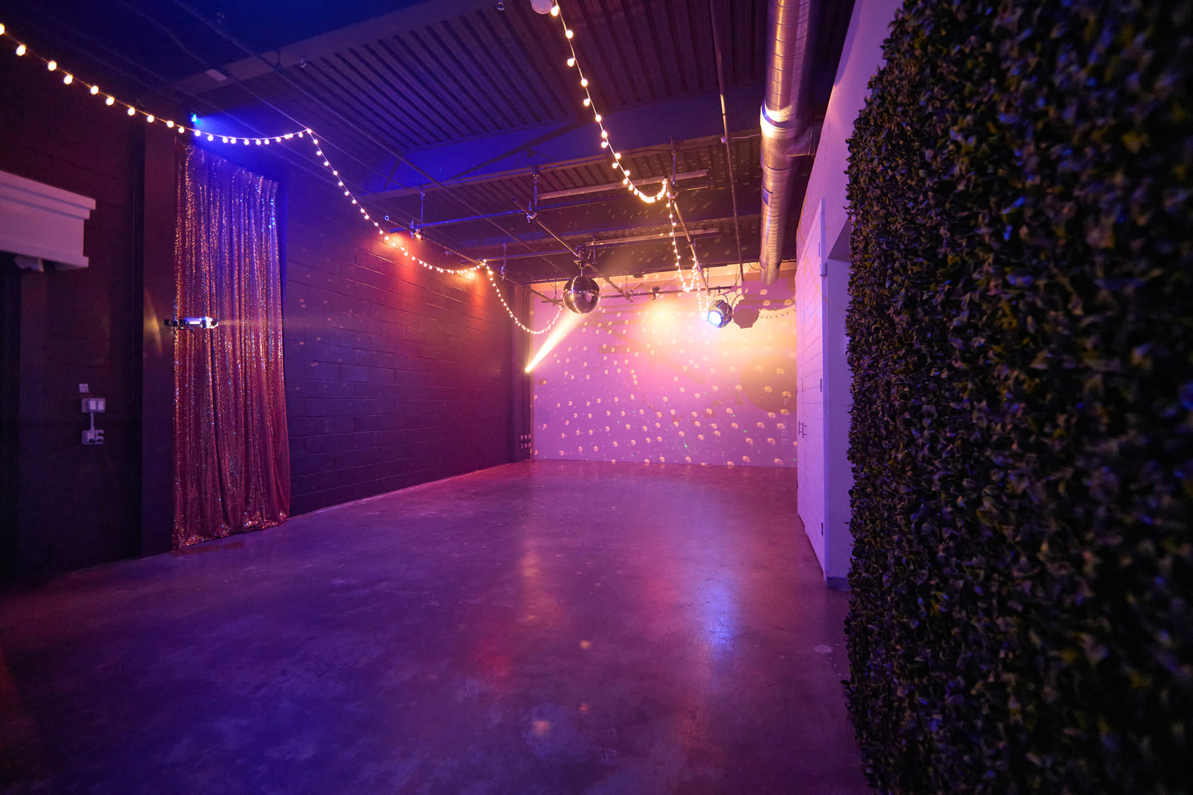 The image shows an empty event space with dim lighting, decorative strings of lights, and a wall covered in greenery.