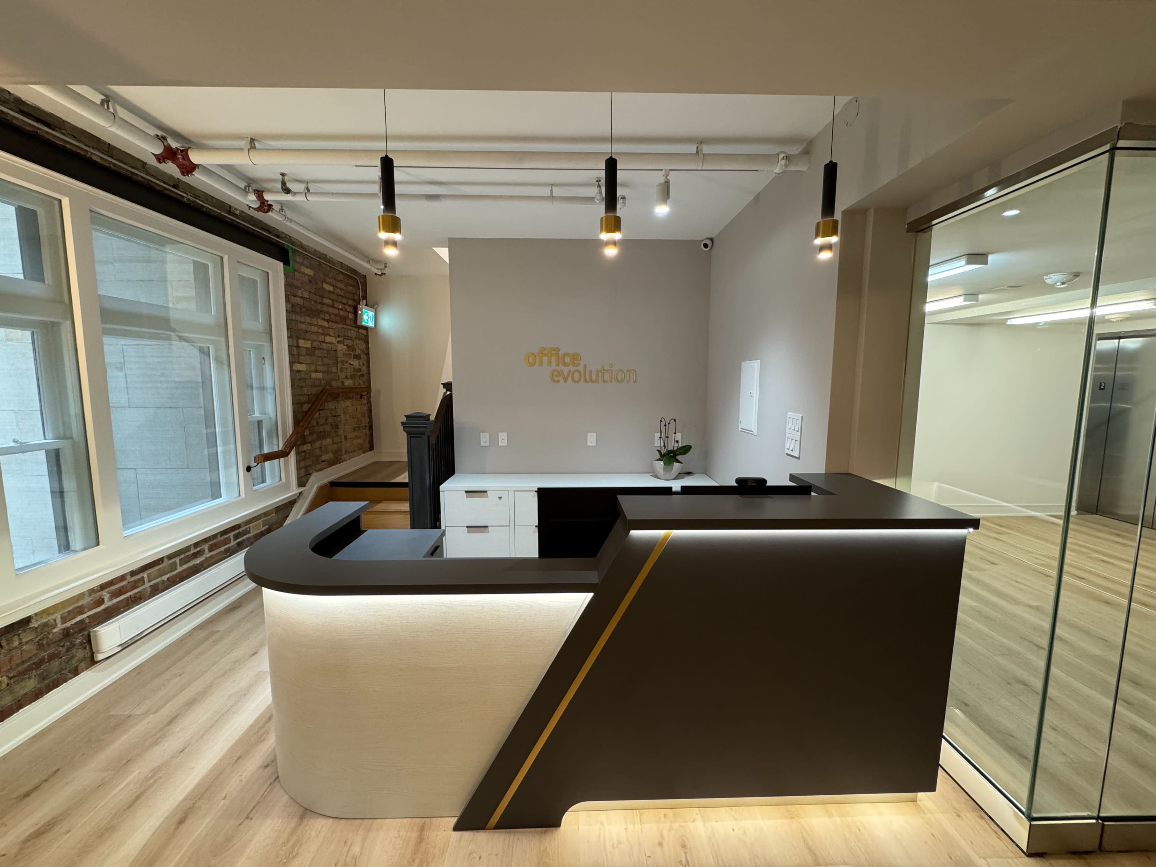 The image shows a modern reception desk with a sleek design, situated in an office space featuring brick walls and large windows.