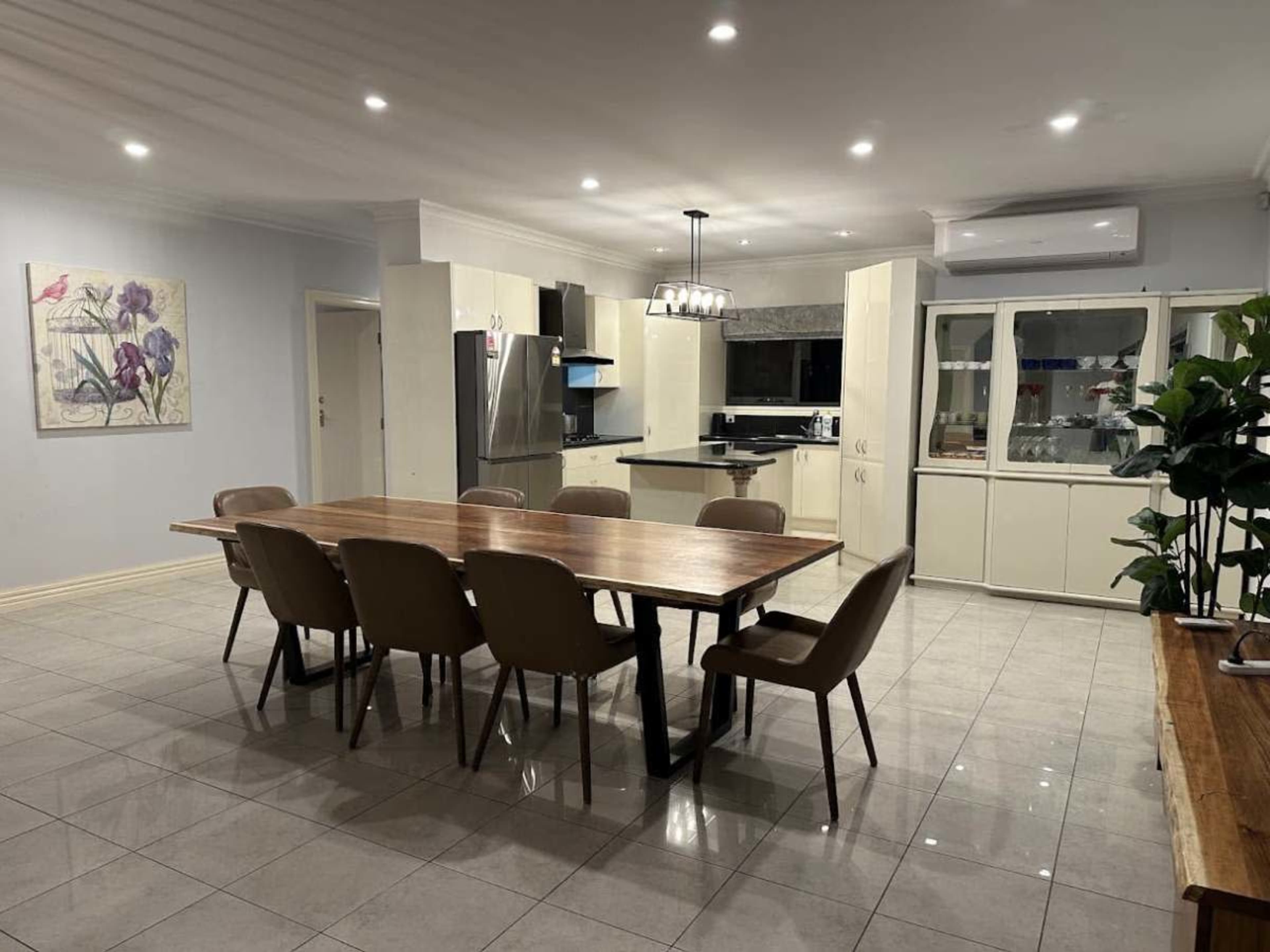 The image shows a spacious dining area with a large wooden table surrounded by eight chairs, adjacent to a modern kitchen with stainless steel appliances and glass-front cabinets.